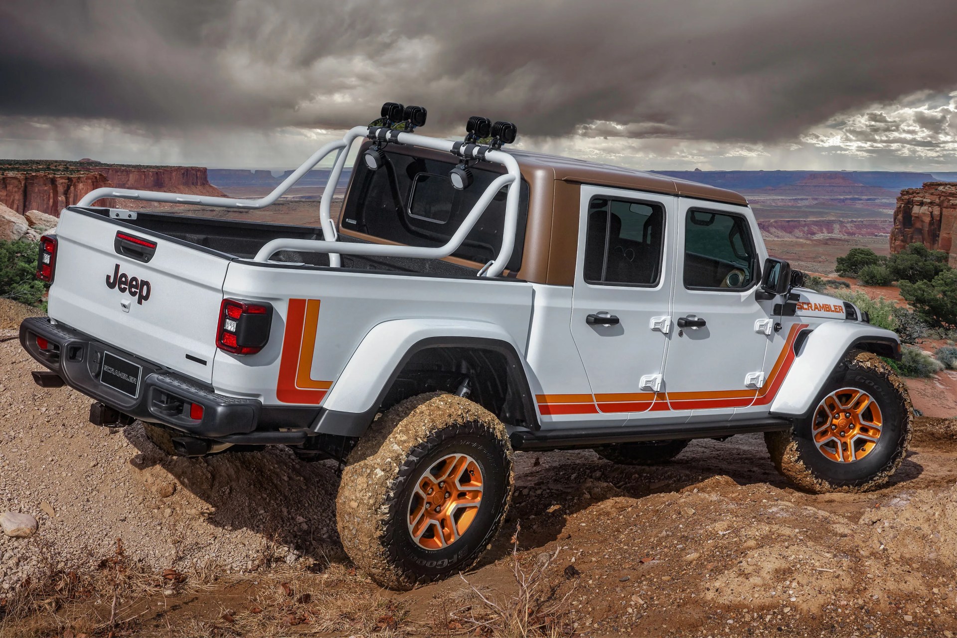 White Jeep Scrambler pickup truck with orange accents and muddy tires on rocky terrain under cloudy sky.