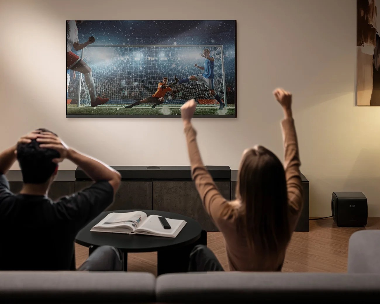 Two people watching a soccer match on a wall-mounted TV with a soundbar and subwoofer in a living room.