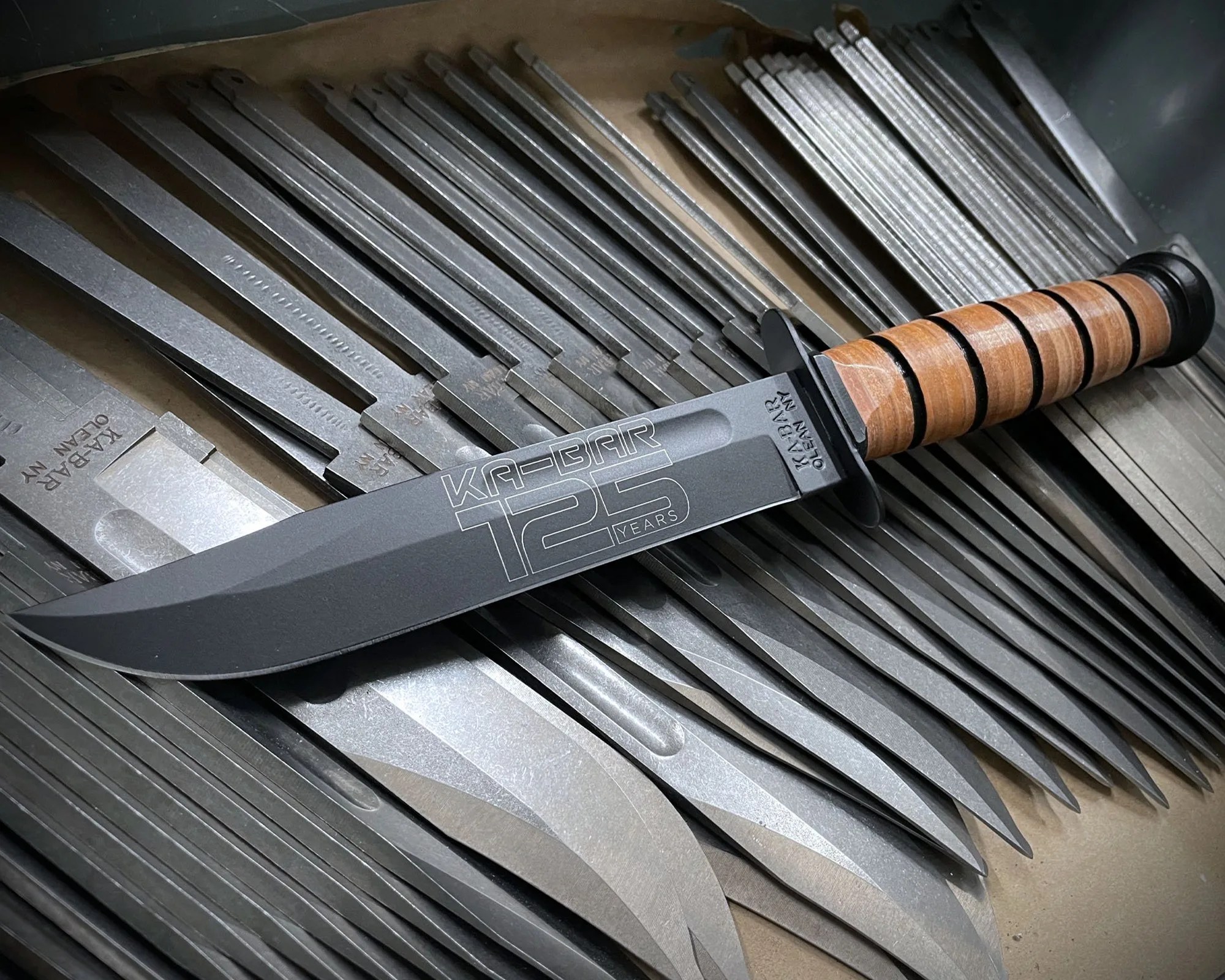 Black KA-BAR 125 Years knife with a wooden handle resting on multiple metal knife blanks.