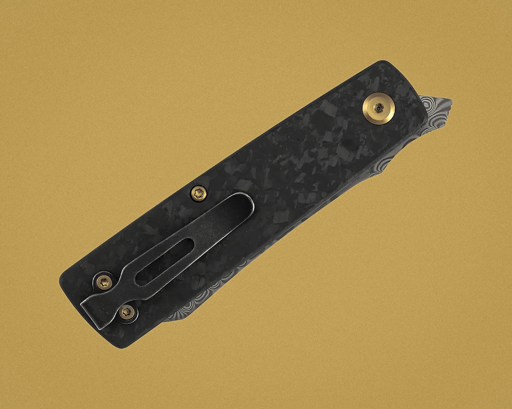 Black textured folding knife handle with brass screws and a partially visible patterned blade on a tan background.