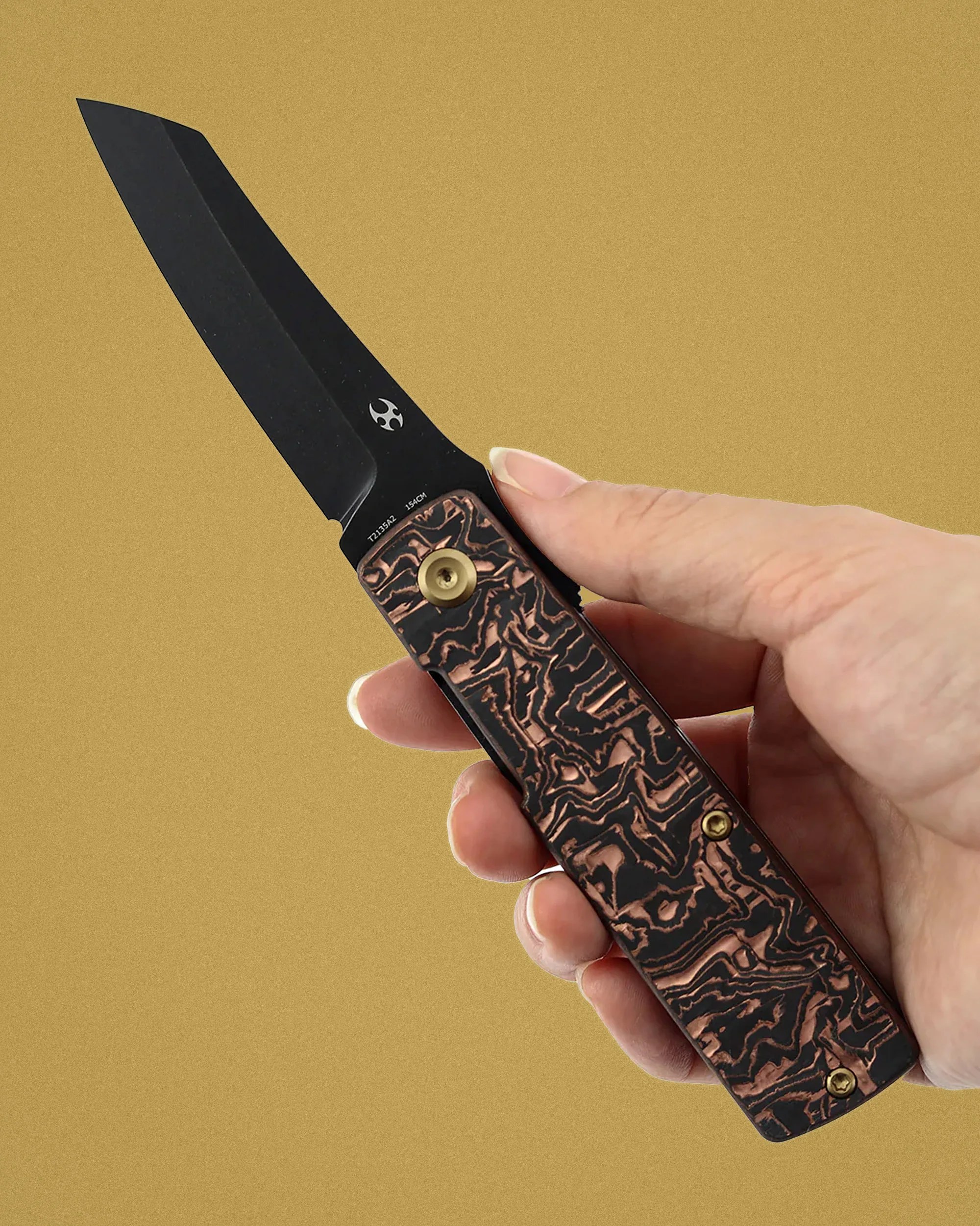 Hand holding a black folding knife with a textured copper and black handle against a tan background.
