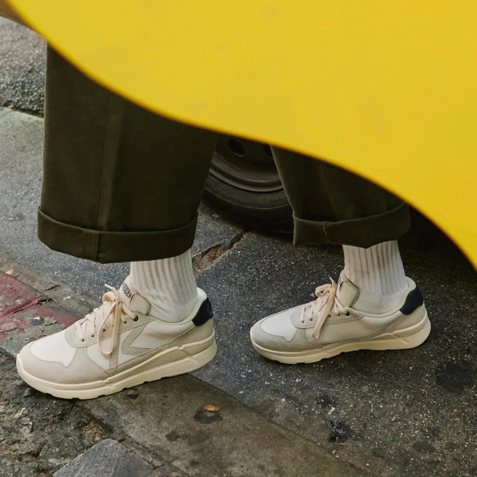White sneakers with beige and black accents worn with white ribbed socks and dark green cuffed pants on pavement.