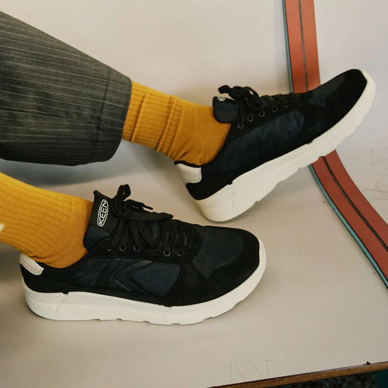 Black and navy KEEN sneakers with white soles worn with mustard yellow socks and gray pinstriped pants.