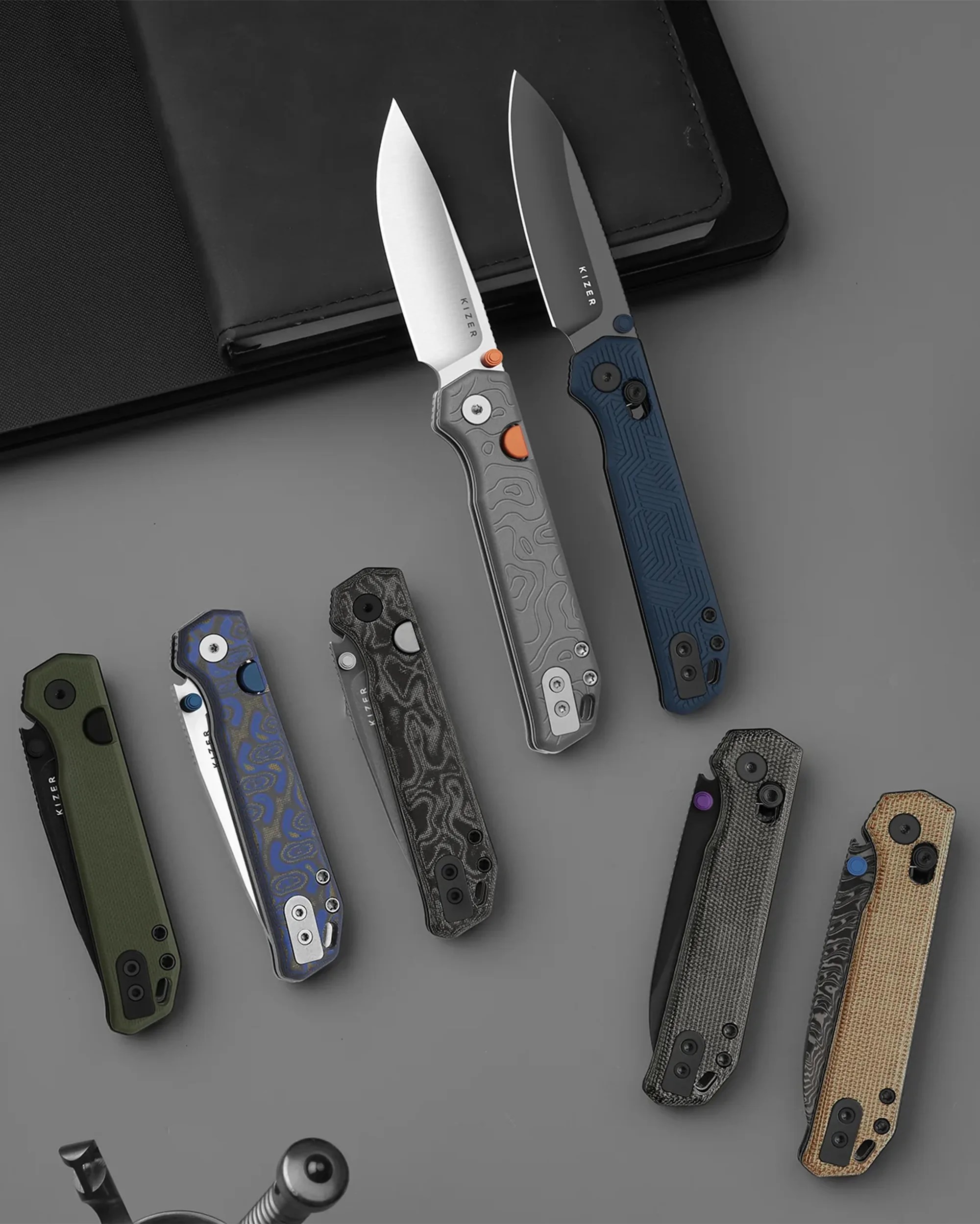 Eight folding knives with patterned handles in various colors, two with blades open, arranged on a gray surface near a black notebook.
