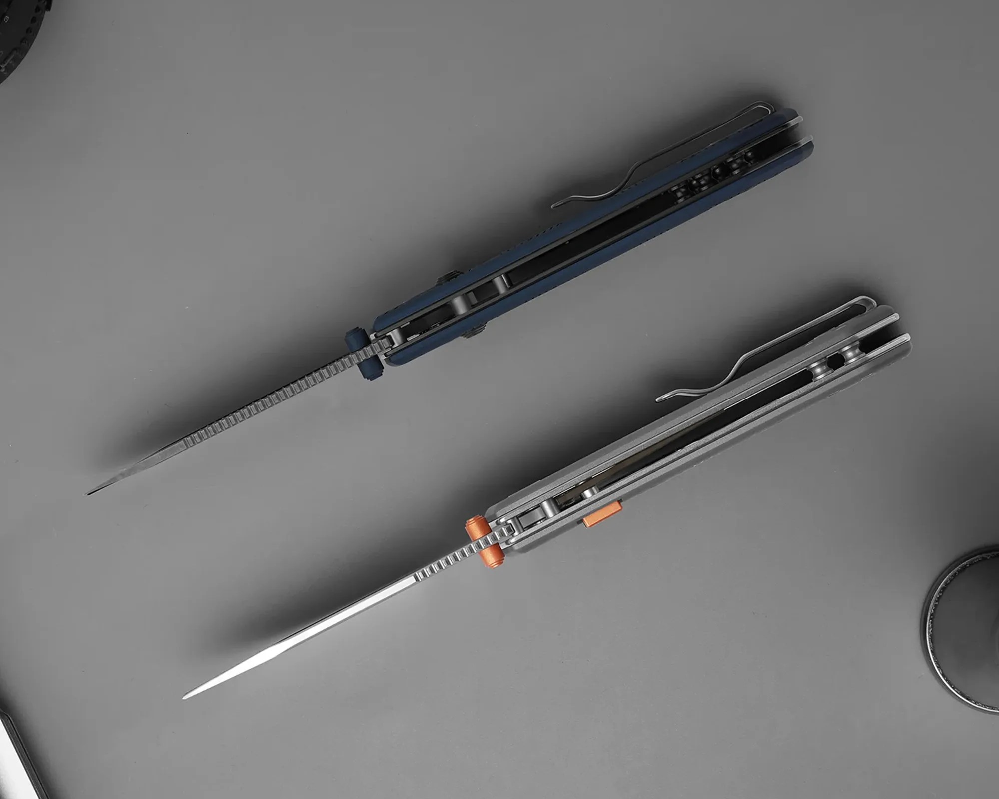 Two folding knives with visible liners and pocket clips, one with a blue handle and the other with a gray handle, on a gray surface.