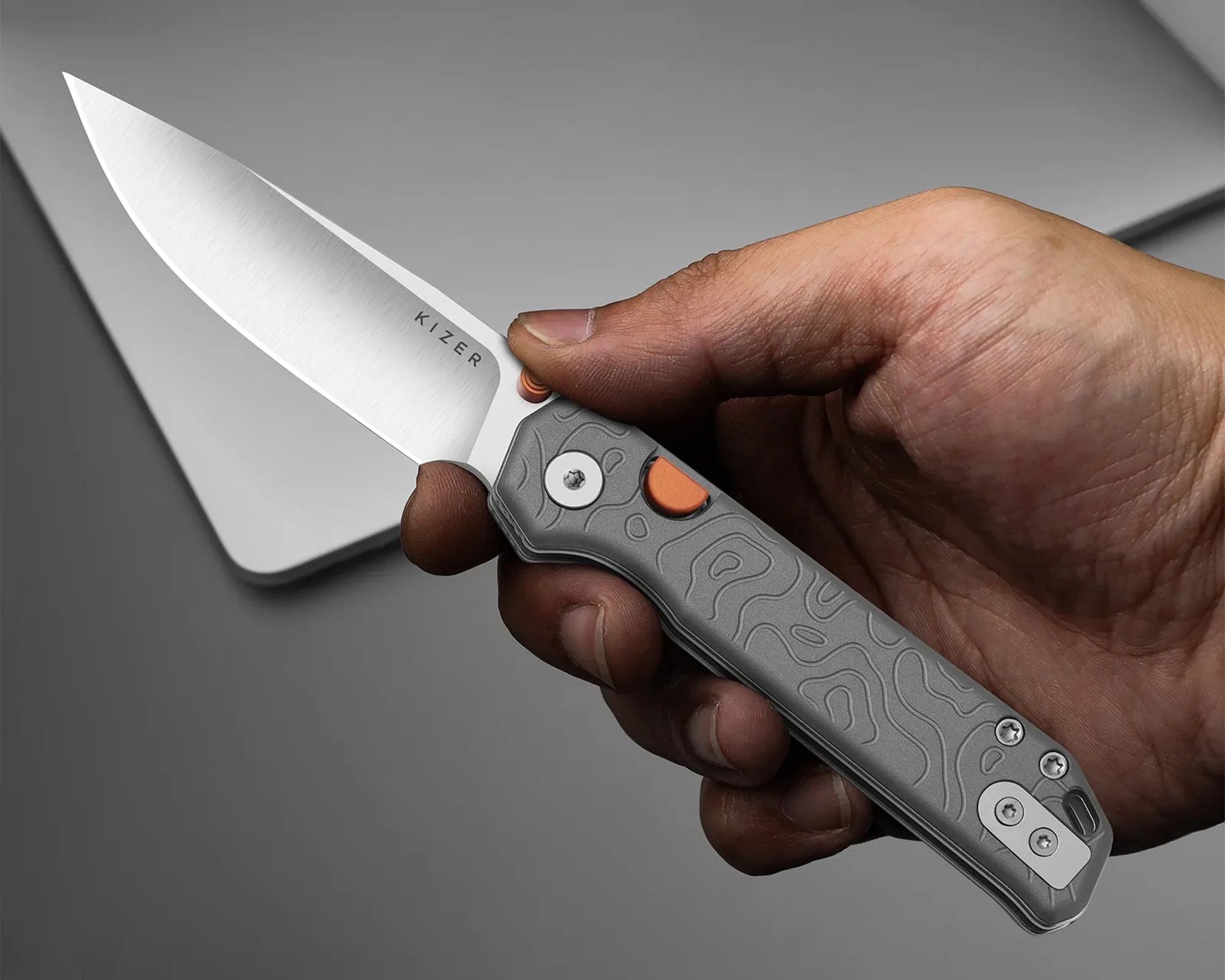 Hand holding a Kizer folding knife with a silver blade and gray textured handle with orange accents.