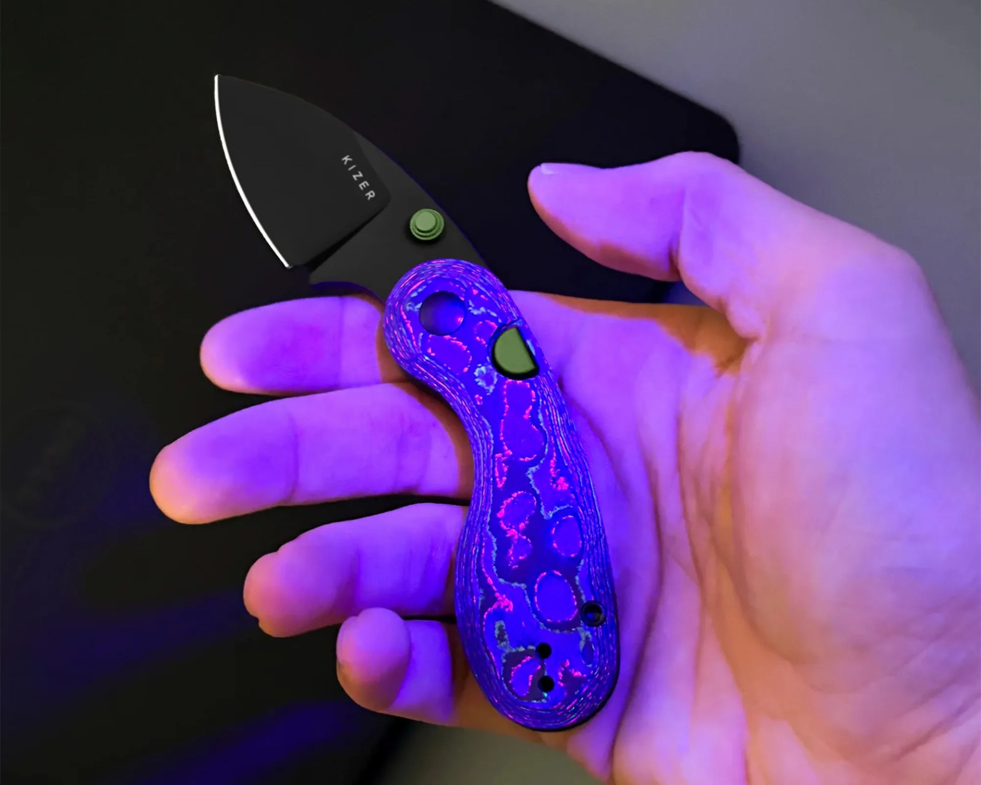 Hand holding a small folding knife with a black blade and a purple patterned handle under blue light.