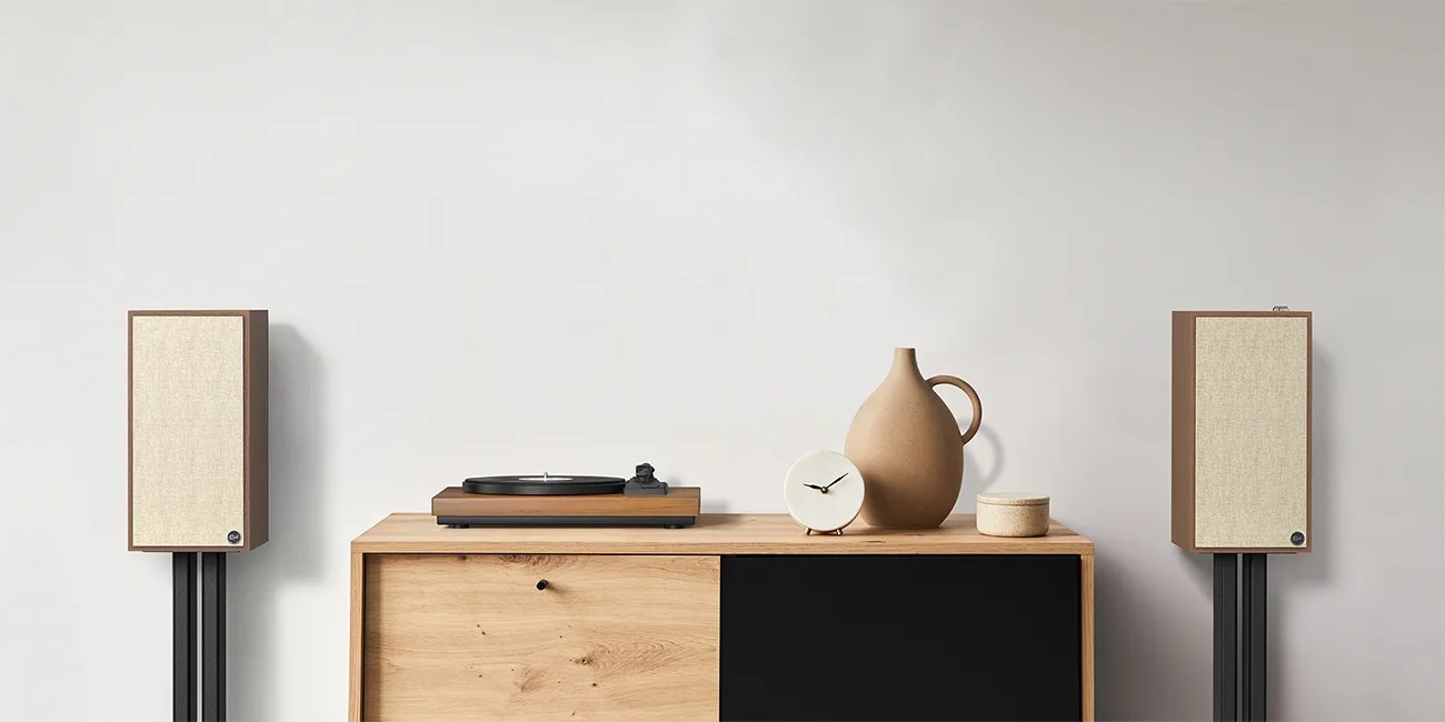 Pair of vintage-style wooden speakers with beige fabric covers on black stands beside a wooden cabinet with a turntable, clock, and pottery.