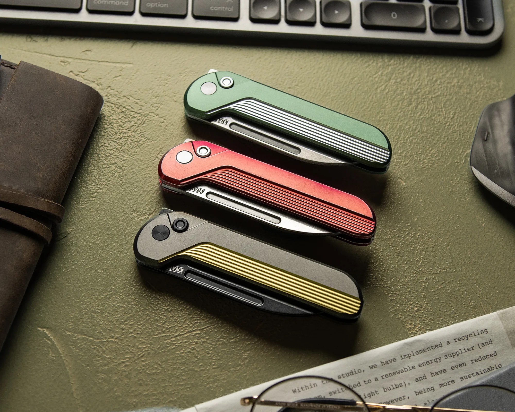 Three closed folding knives with ribbed handles in green, red, and gray with yellow accents on a textured green surface near a keyboard, leather item, and paper with glasses.
