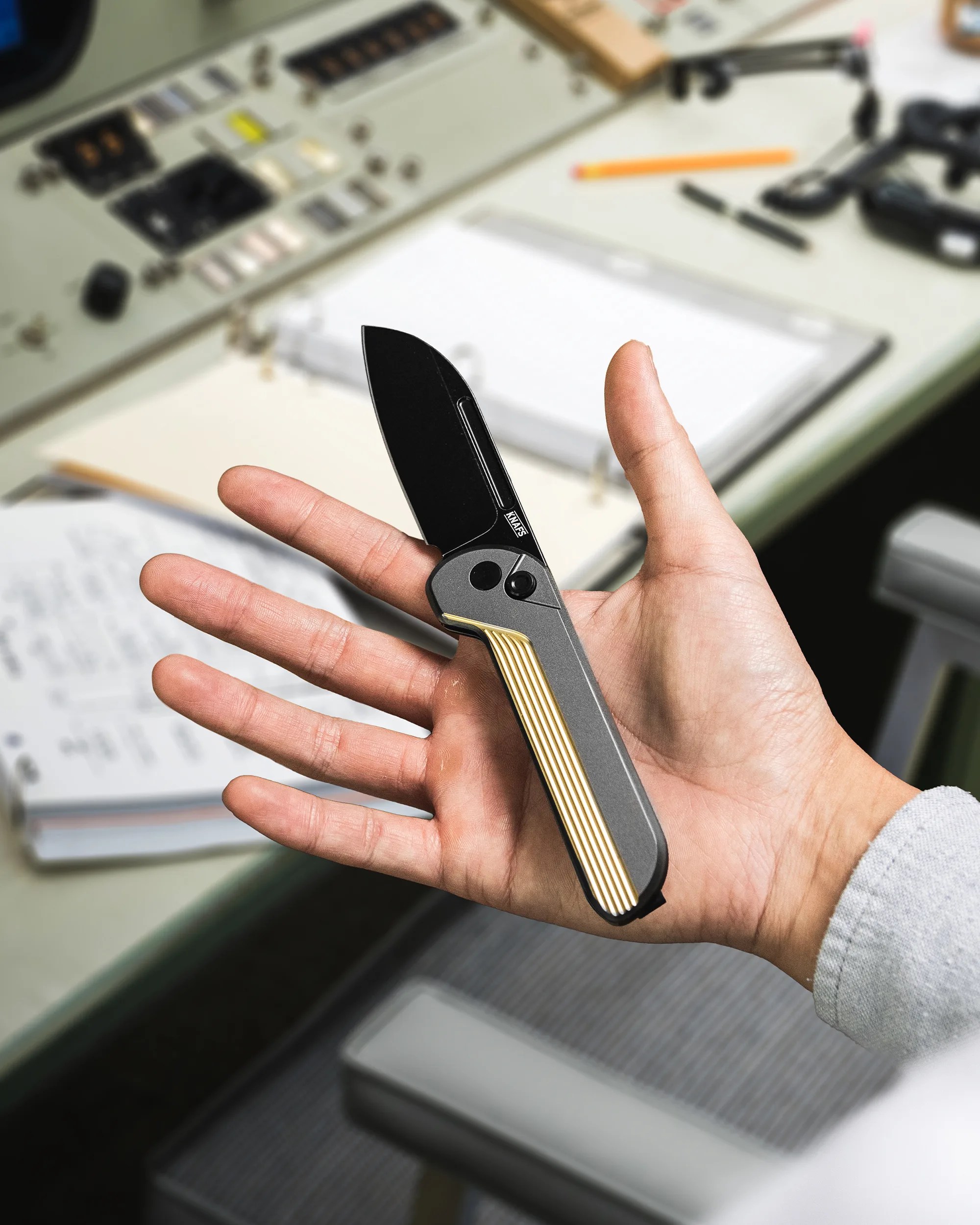 Hand holding a folding knife with a black blade and gray handle featuring gold stripes.