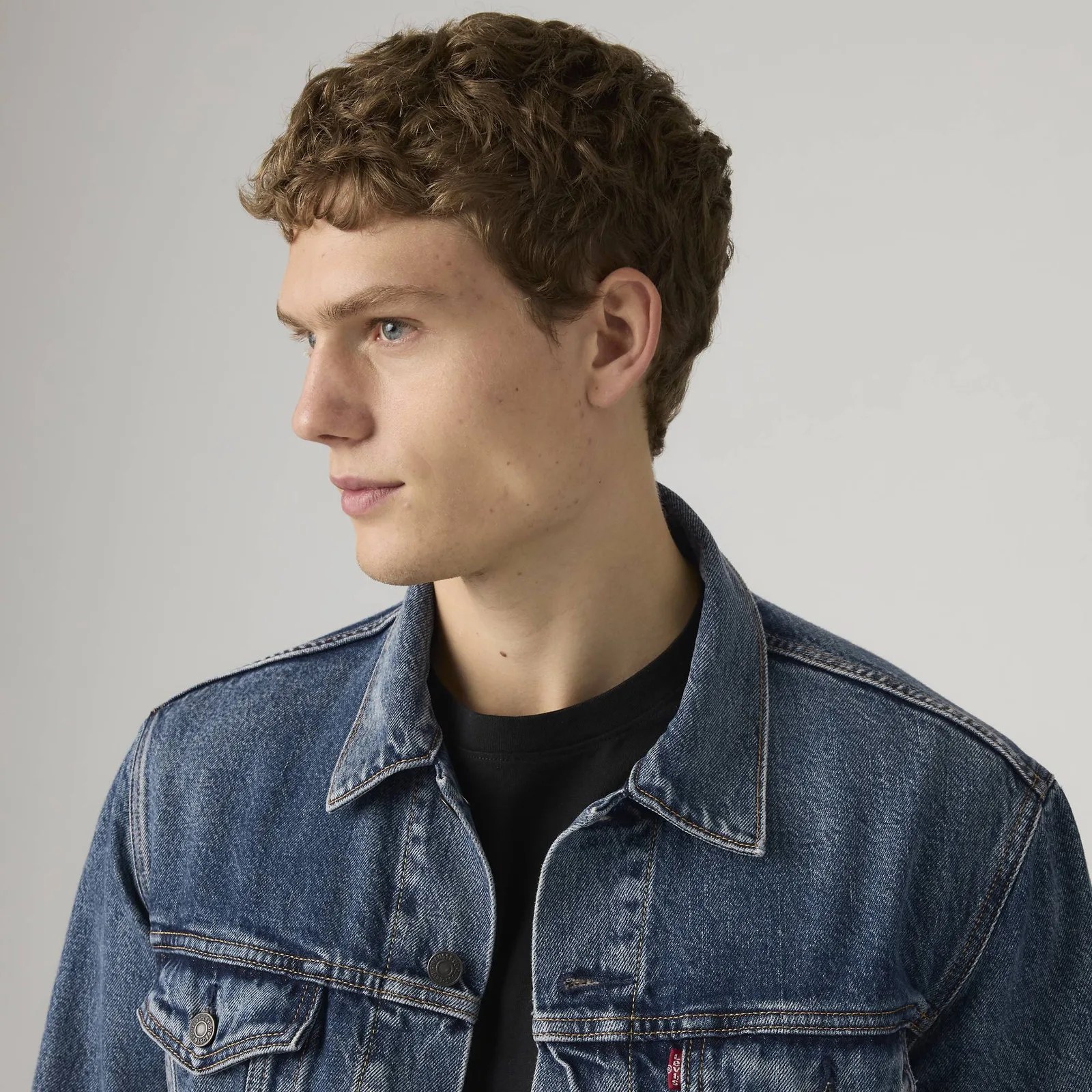 Young person with short curly brown hair wearing a blue denim jacket over a black shirt, facing left.
