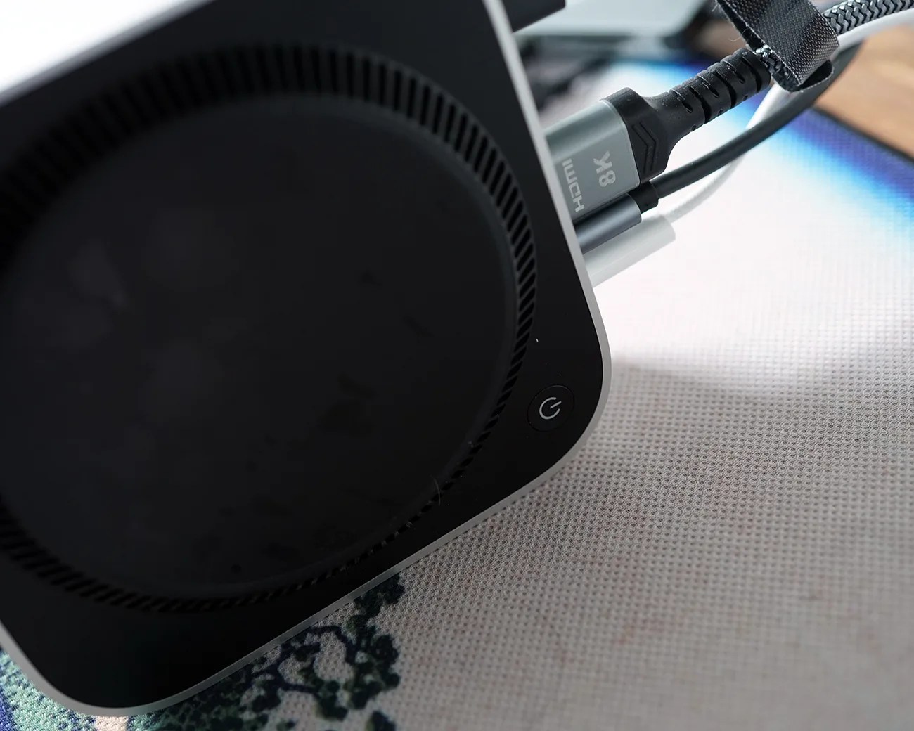 Close-up of a black and silver electronic device with an HDMI 8K cable plugged in, resting on a patterned fabric surface.
