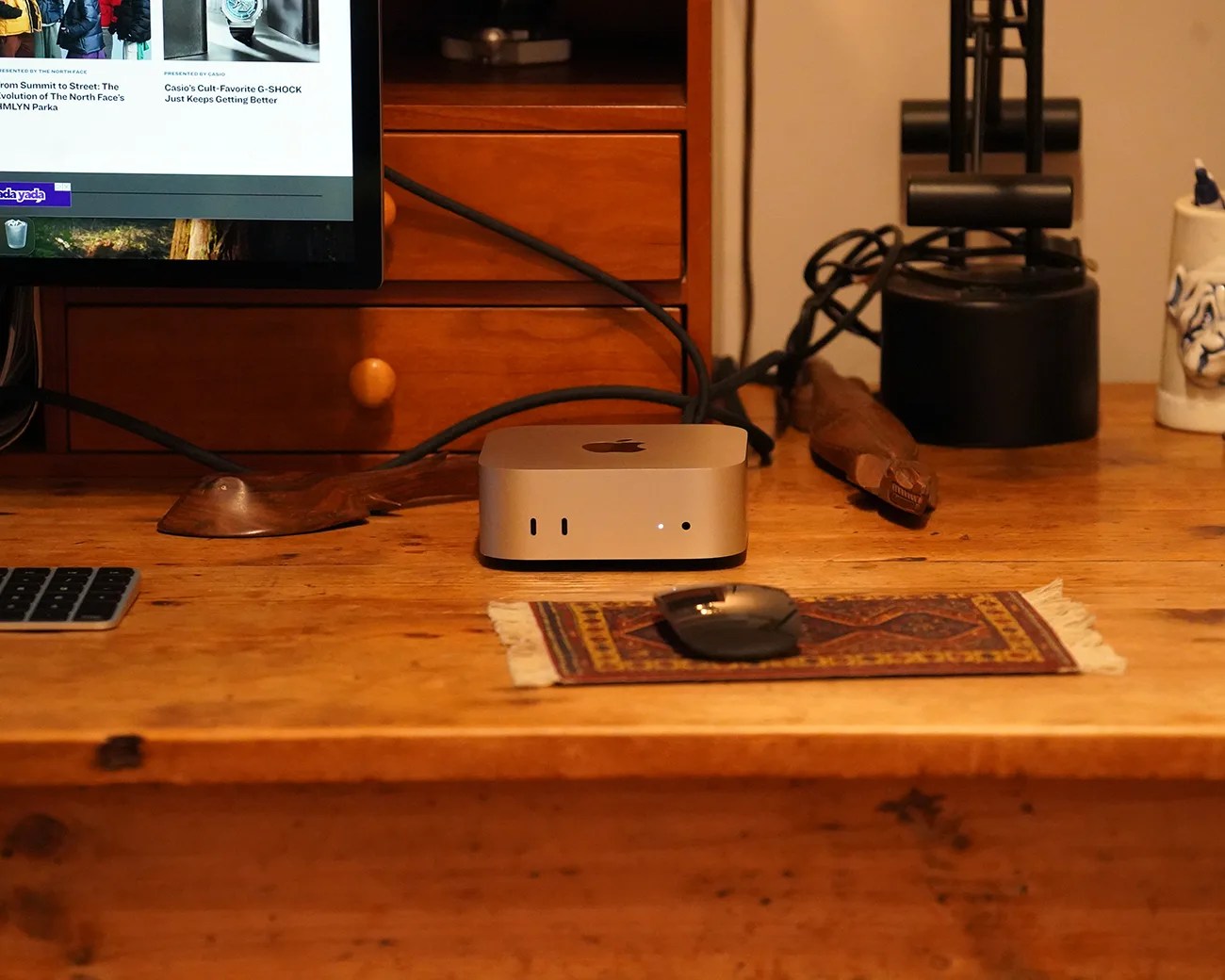Silver Apple Mac Mini on a wooden desk with a black mouse on a small patterned rug and a monitor partially visible.