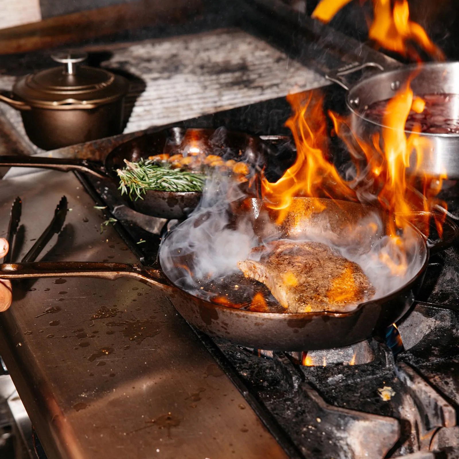 Cast iron skillet with steak cooking over open flame on a gas stove, with another skillet holding rosemary and garlic.