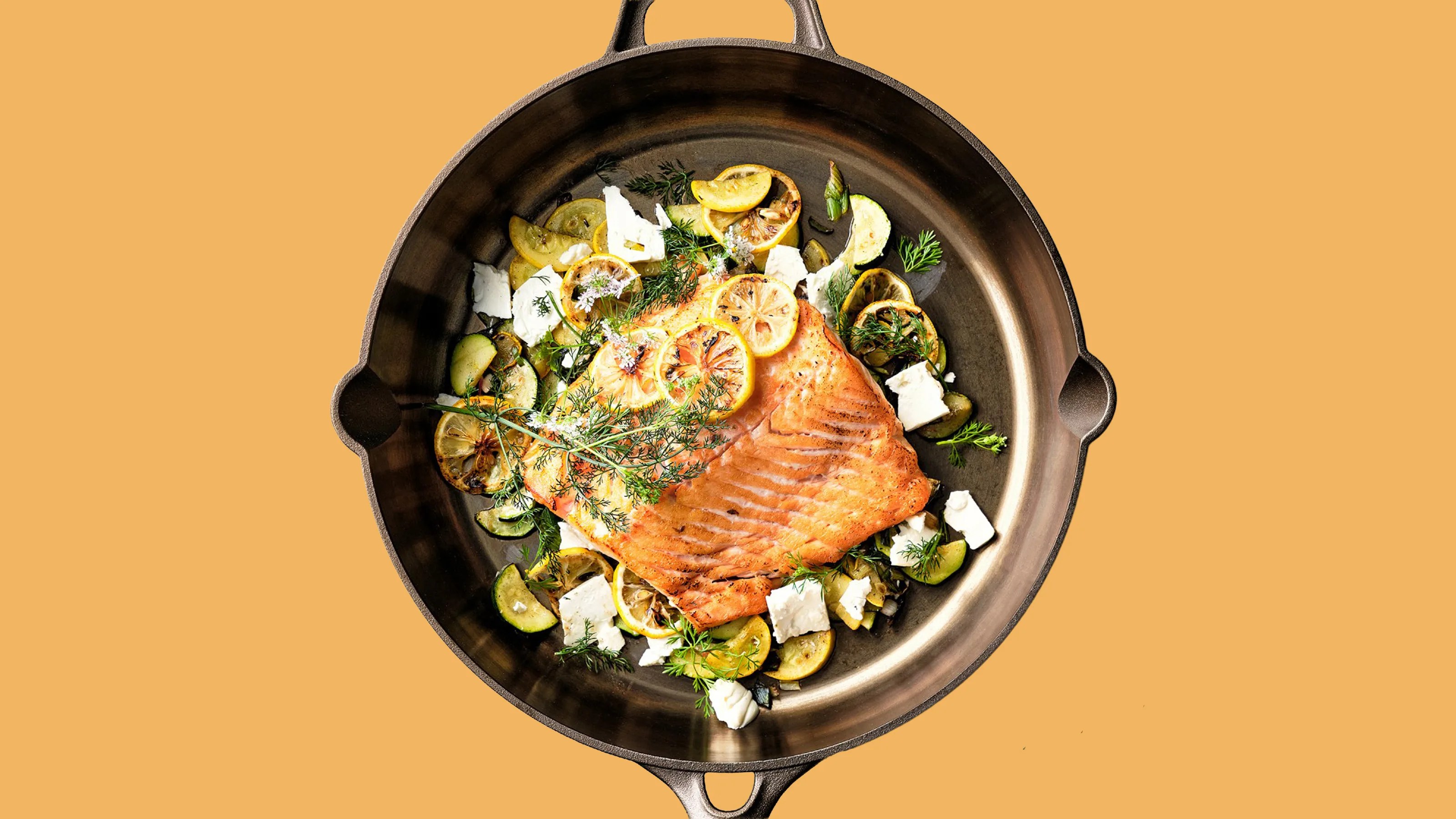 Cast iron skillet with cooked salmon topped with lemon slices, herbs, zucchini, and white cheese chunks.