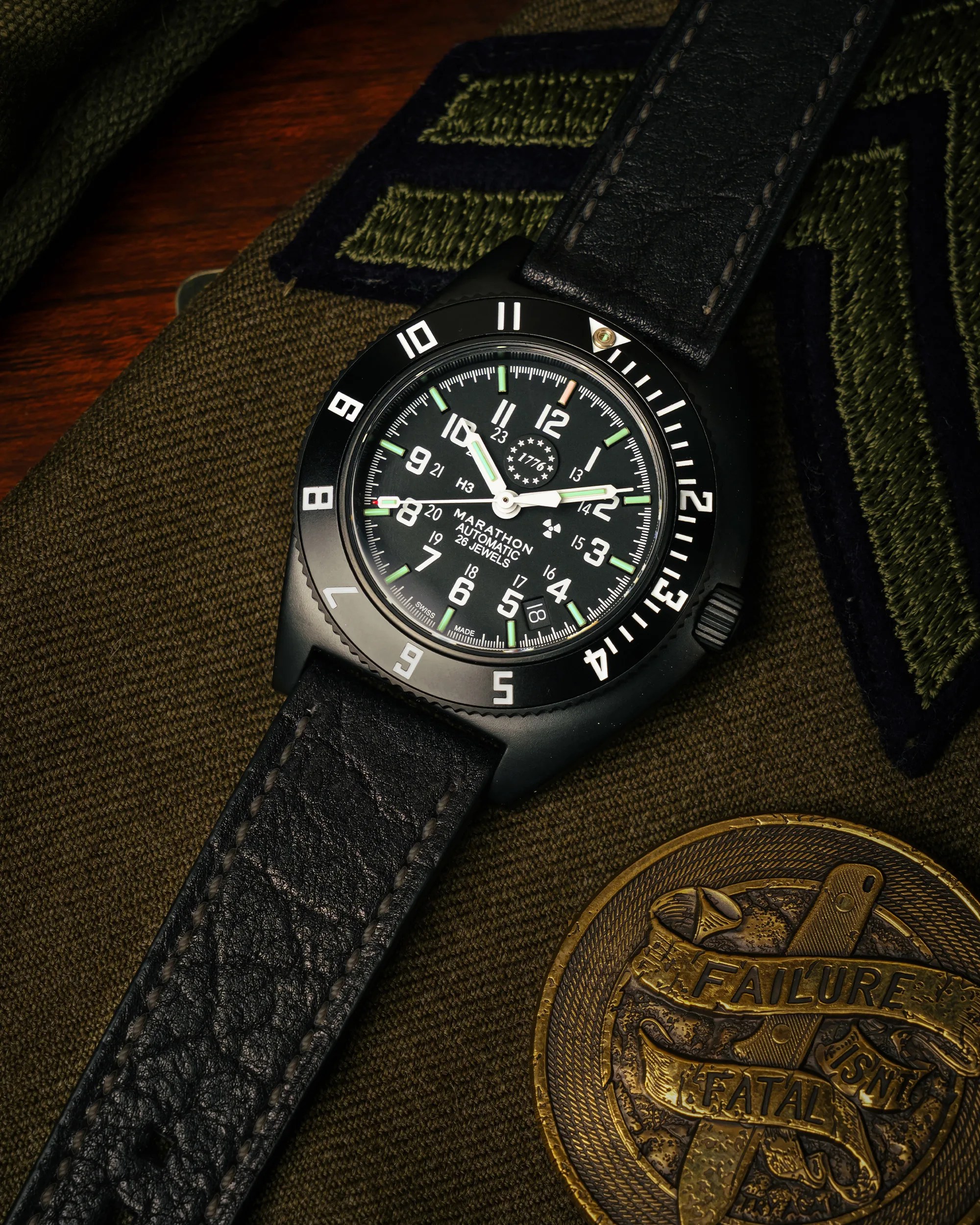 Black Marathon automatic wristwatch with white numerals and green luminous markers on a black leather strap.