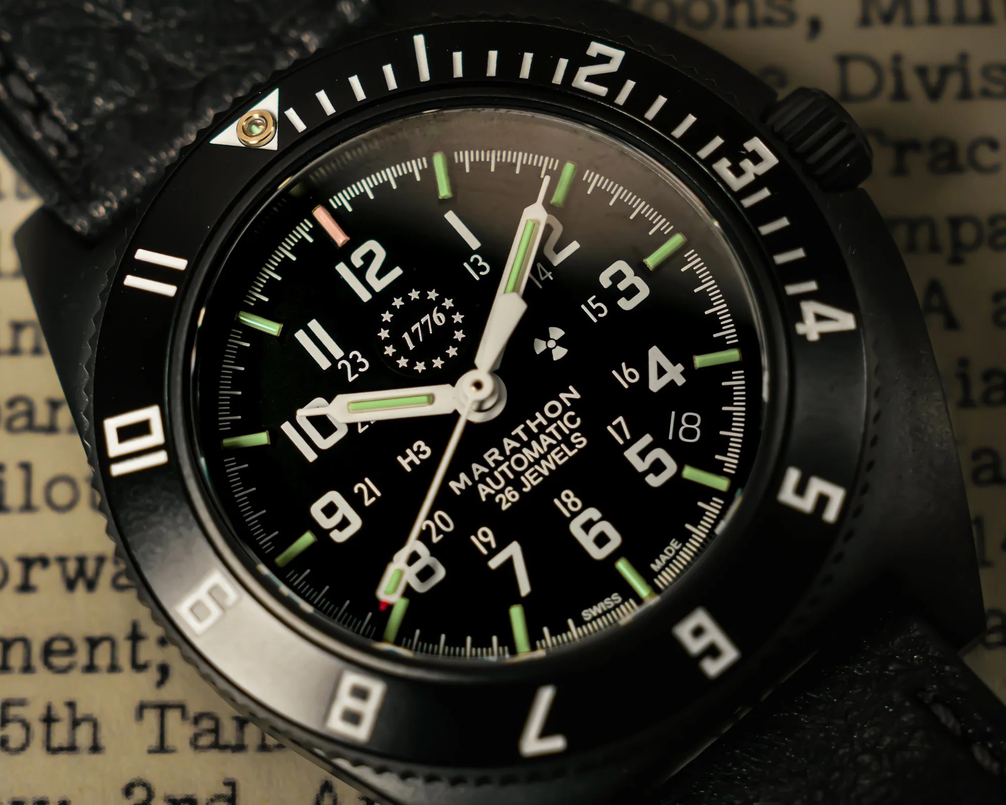 Close-up of a black Marathon automatic wristwatch with white numerals, green luminous hands, and a black bezel on a textured black strap.