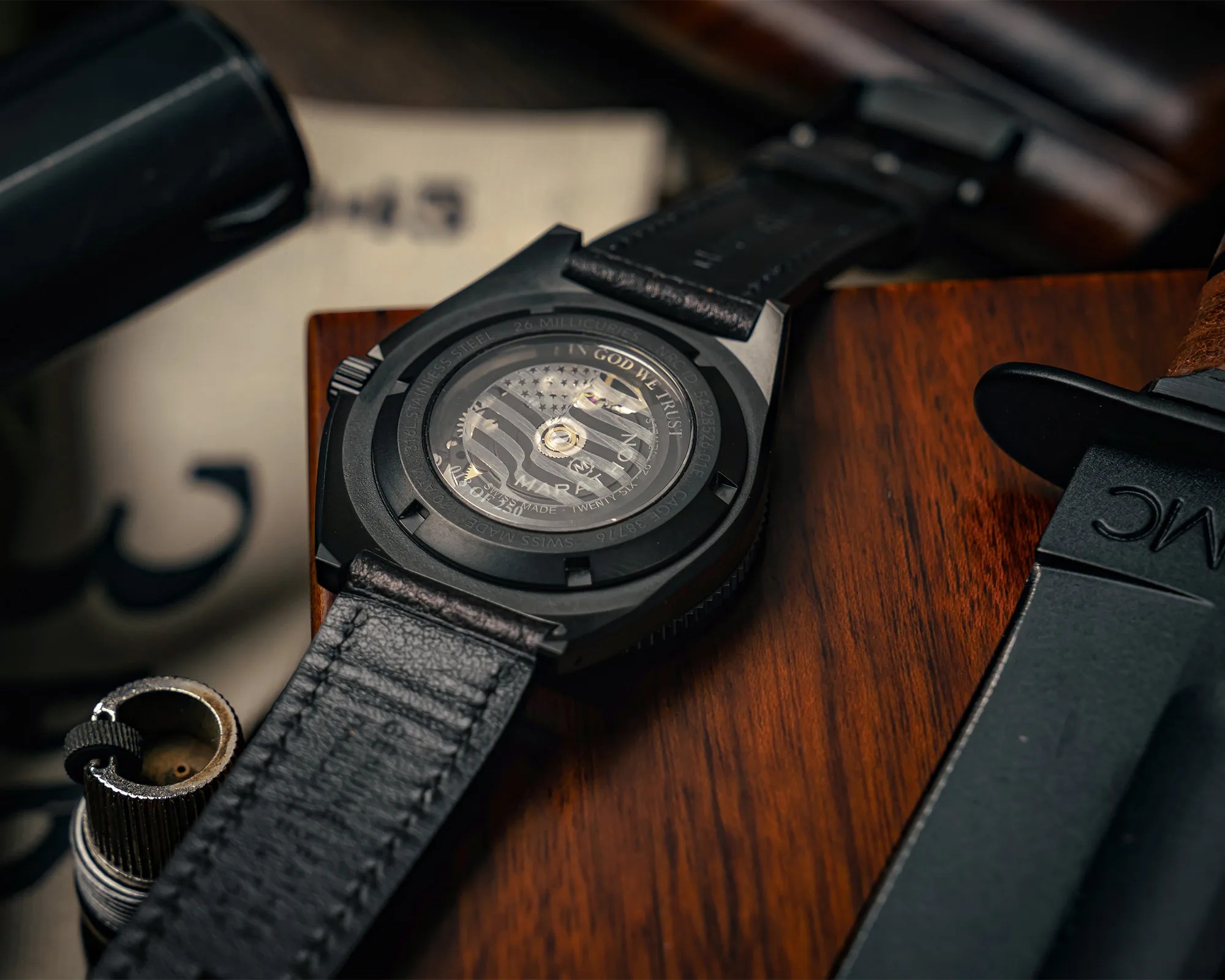 Close-up of a black wristwatch with a transparent back showing internal gears and "IN GOD WE TRUST" text, on a wooden surface.