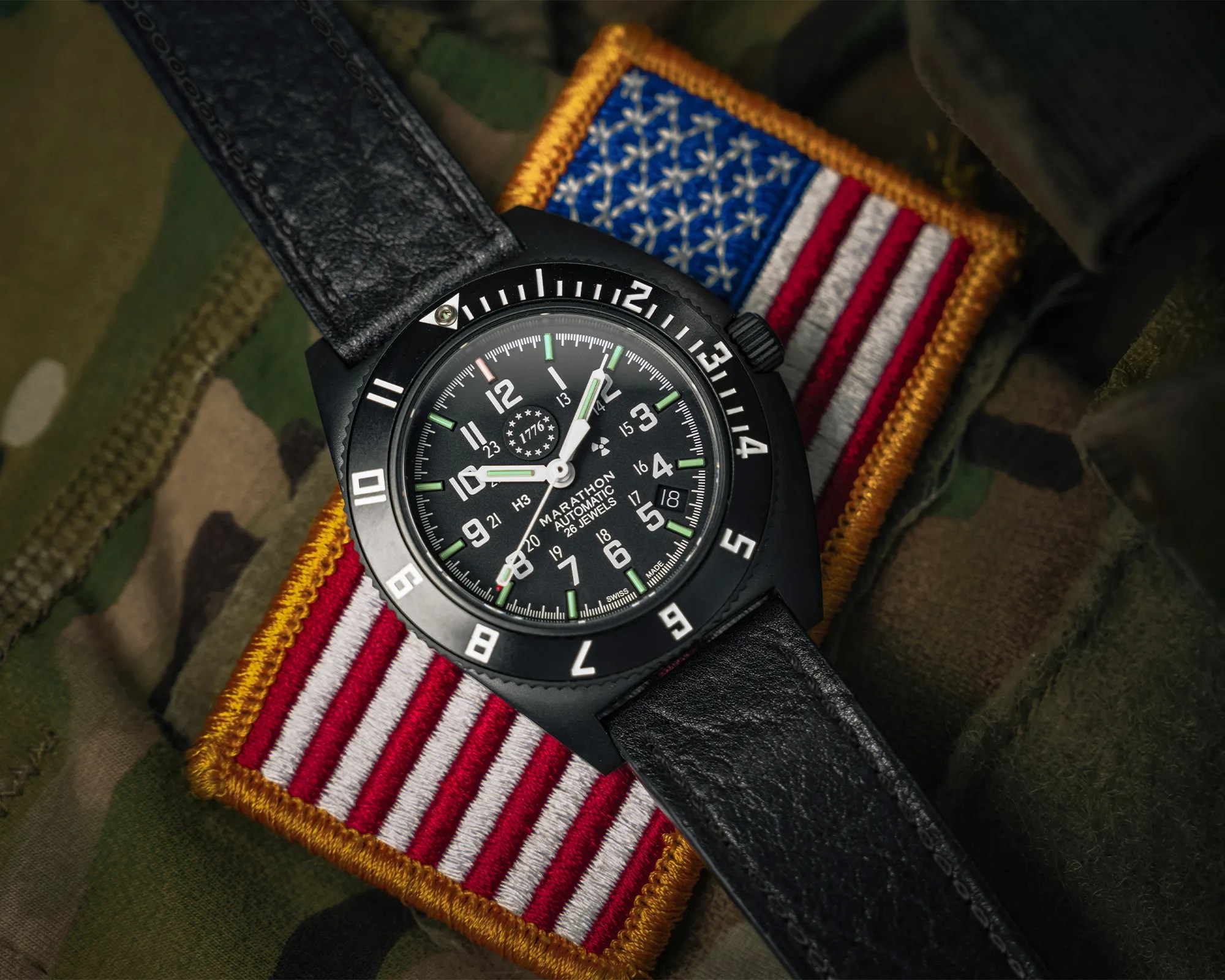 Black Marathon automatic wristwatch with luminous hands and markers on a black leather strap over a U.S. flag patch on camouflage fabric.