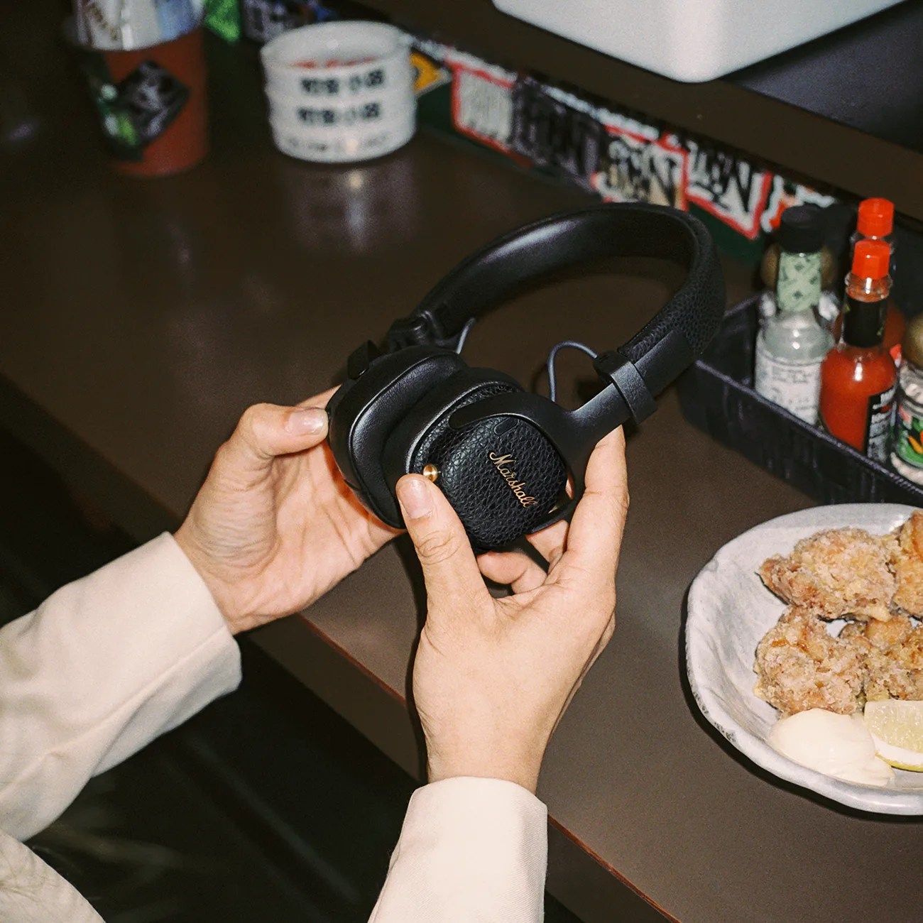 Hands holding black Marshall over-ear headphones above a brown table with a plate of fried chicken and condiments.