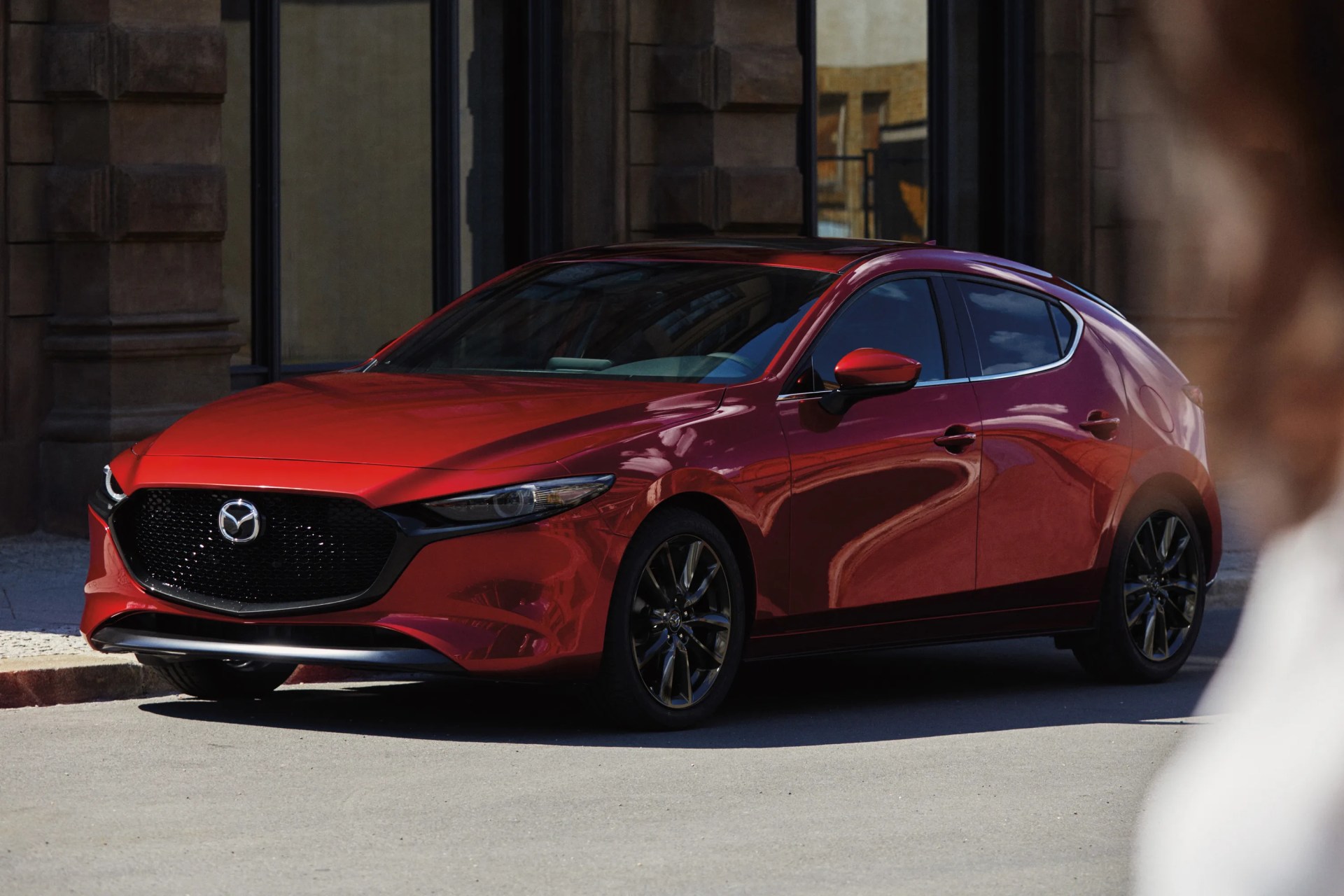 Red Mazda hatchback with black grille and dark alloy wheels parked on a street near a stone building.