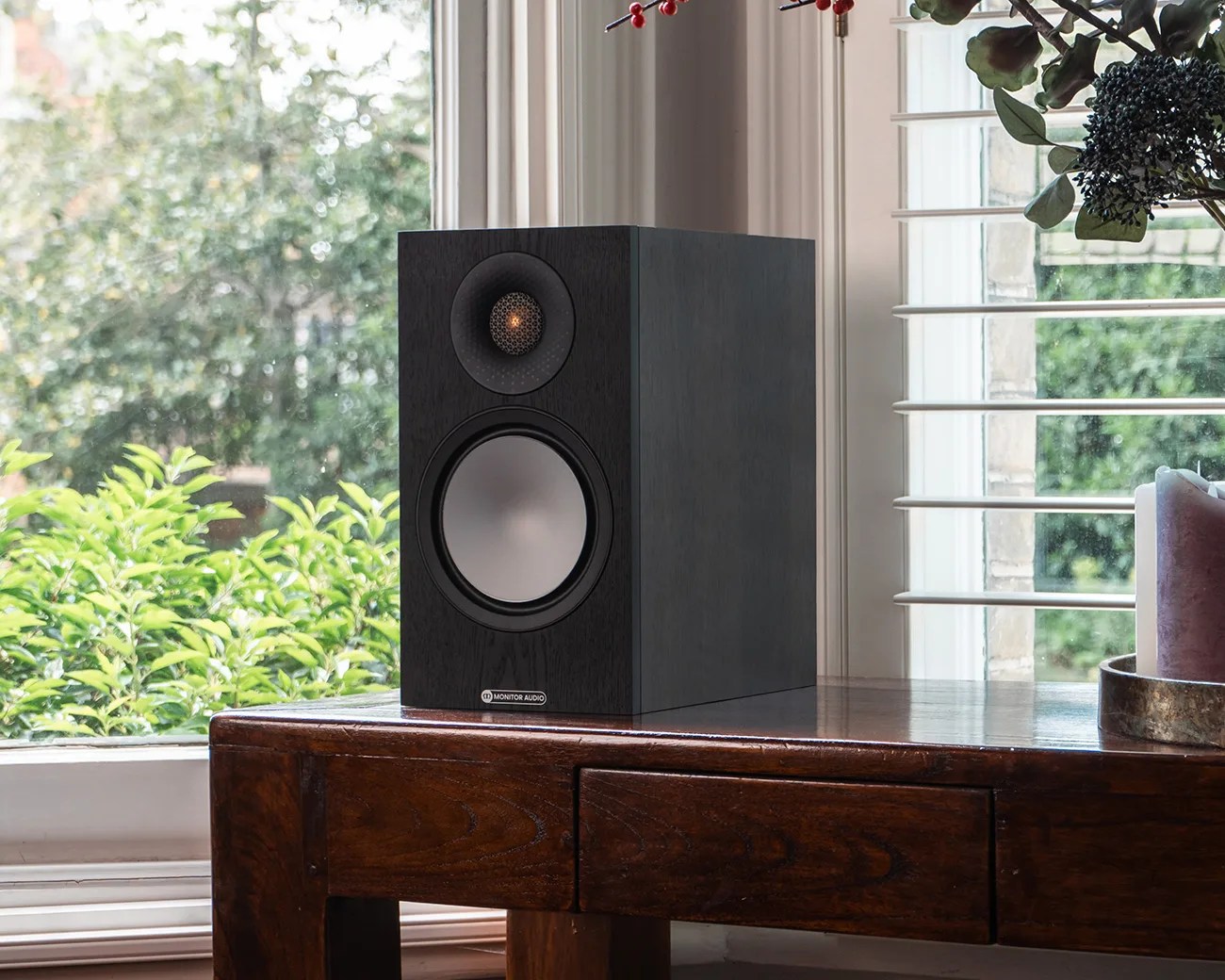 Black Monitor Audio speaker with silver woofer on a wooden table near a window with greenery outside.