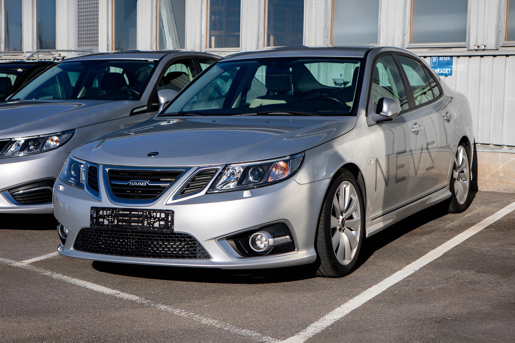 Silver Saab sedan parked in a marked parking space in front of a building with large windows.