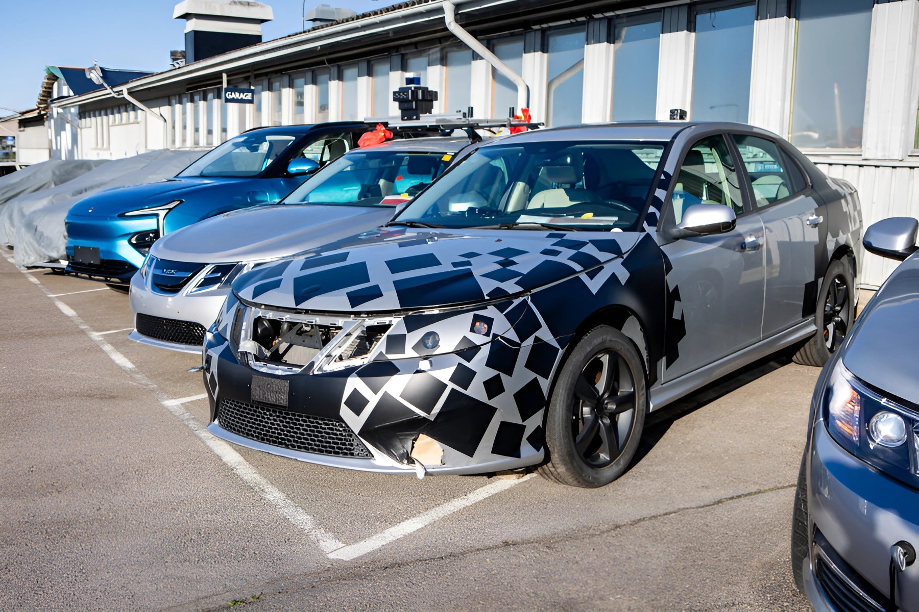 Silver sedan with black and silver camouflage wrap on front bumper and hood parked next to other cars in a lot.
