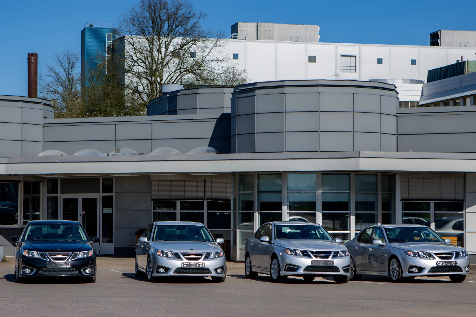 Four Saab sedans, three silver and one black, parked in front of a modern gray building with large windows.
