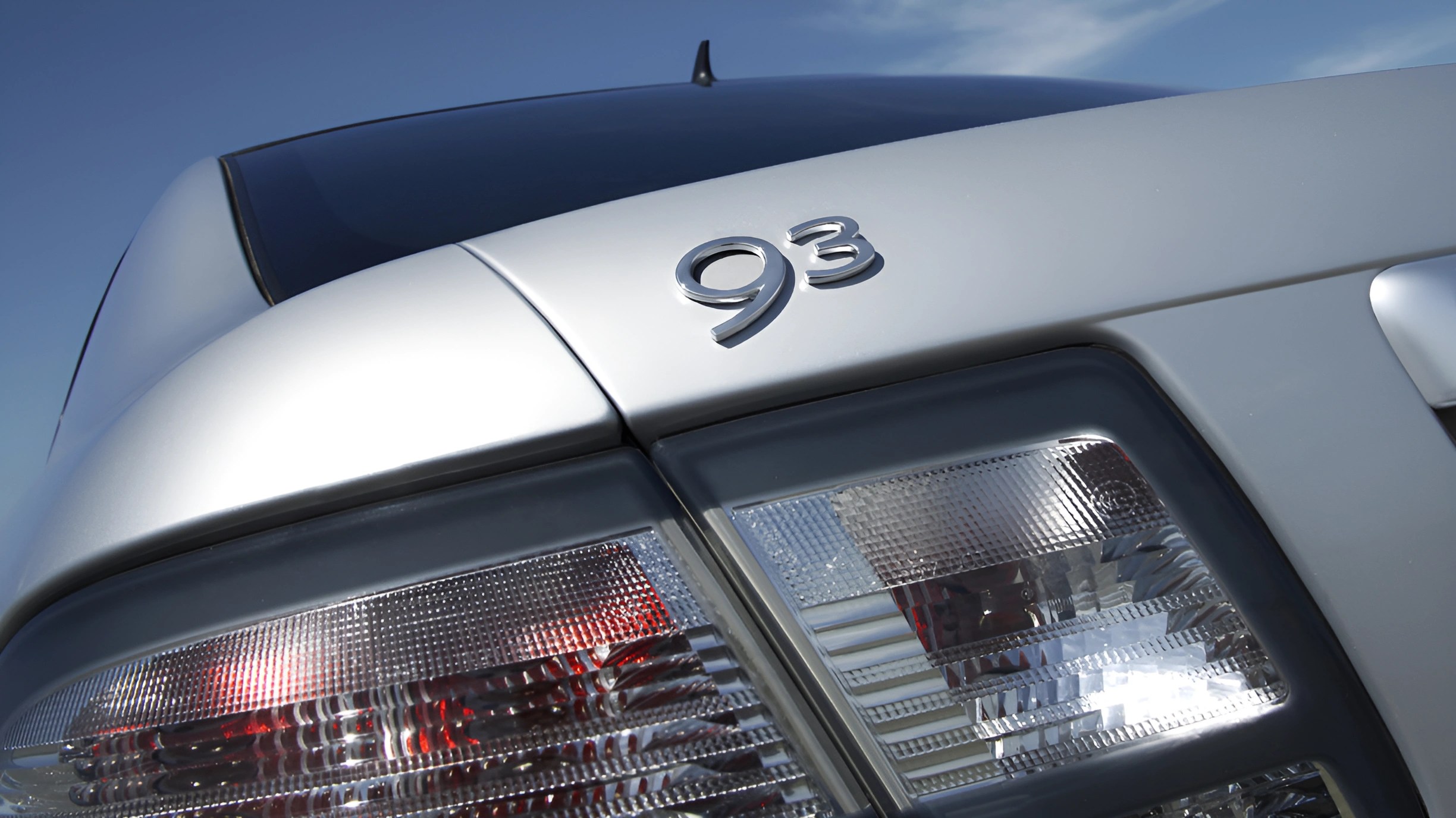 Close-up of silver car rear with "93" badge and clear taillights under blue sky.