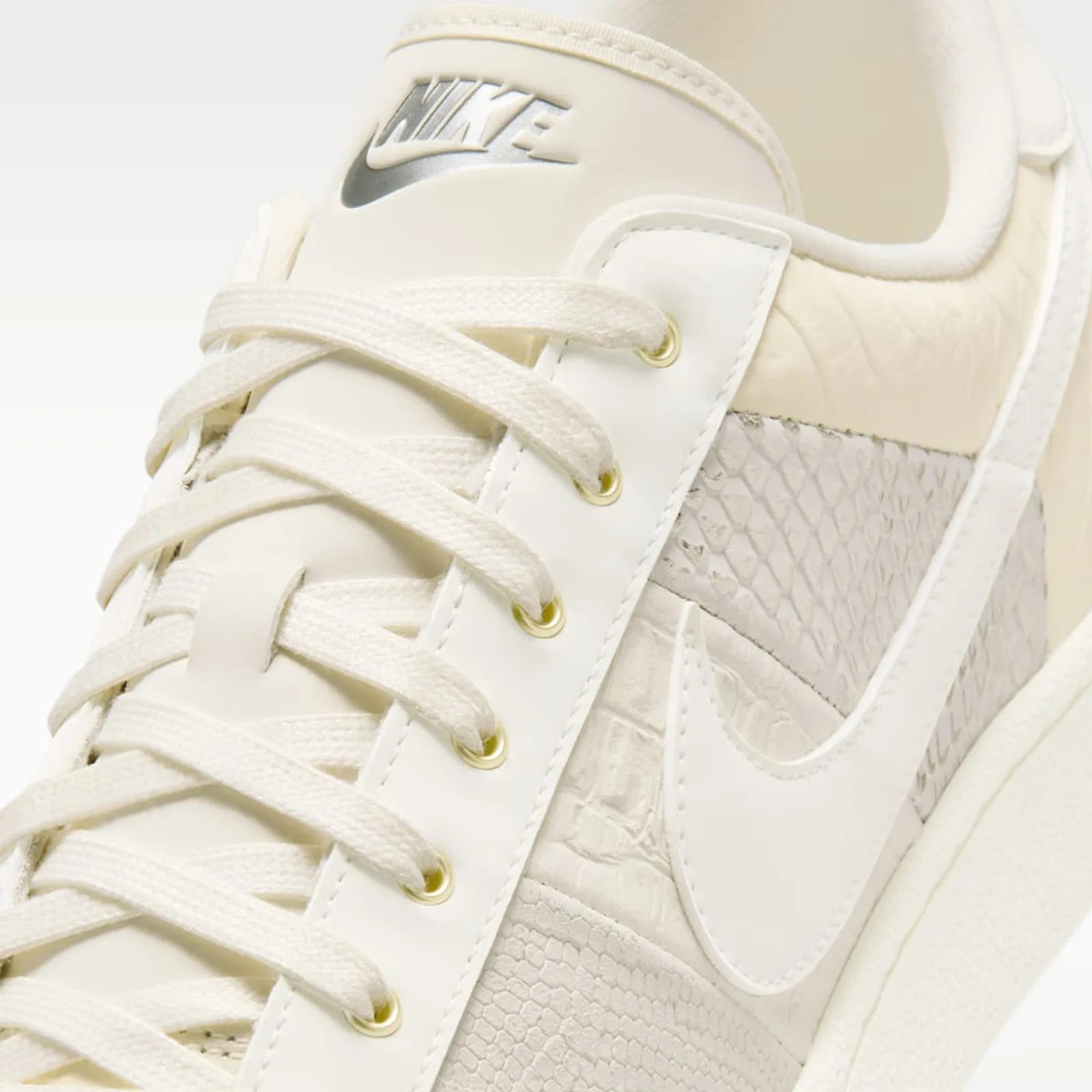 Close-up of a cream Nike sneaker with textured snake-skin pattern and cream laces threaded through gold eyelets.