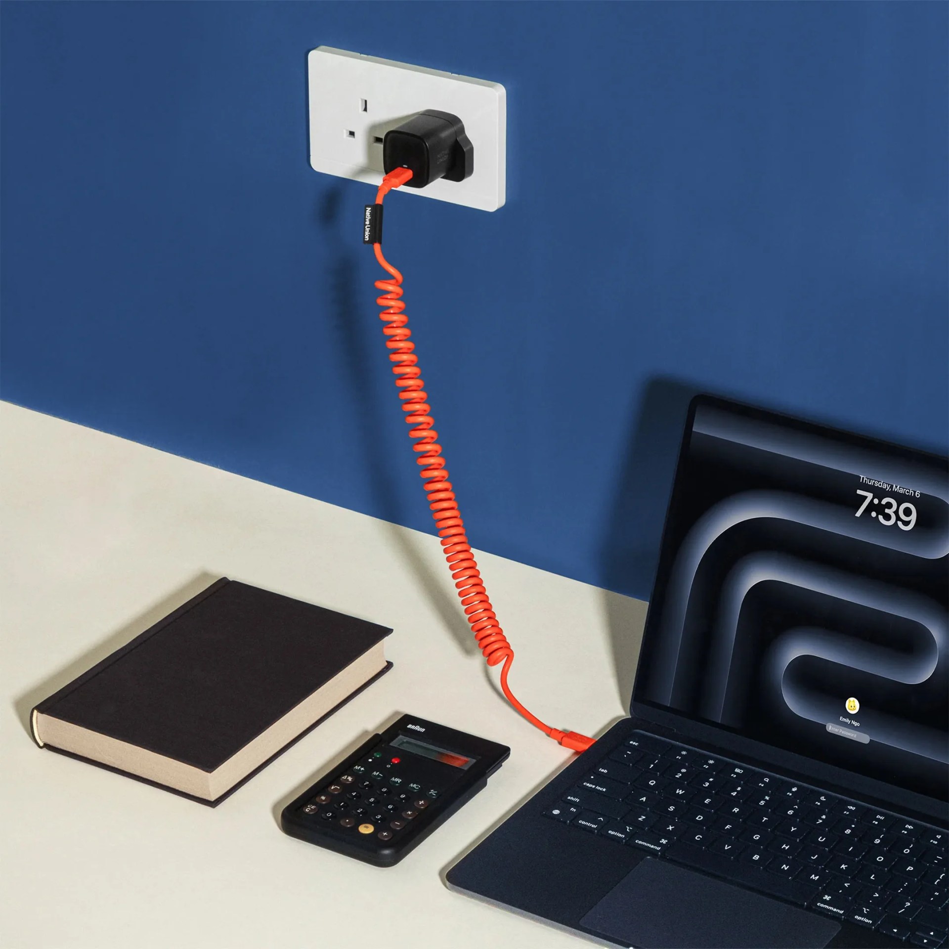 Black laptop with backlit keyboard charging via orange coiled cable plugged into a white wall socket.