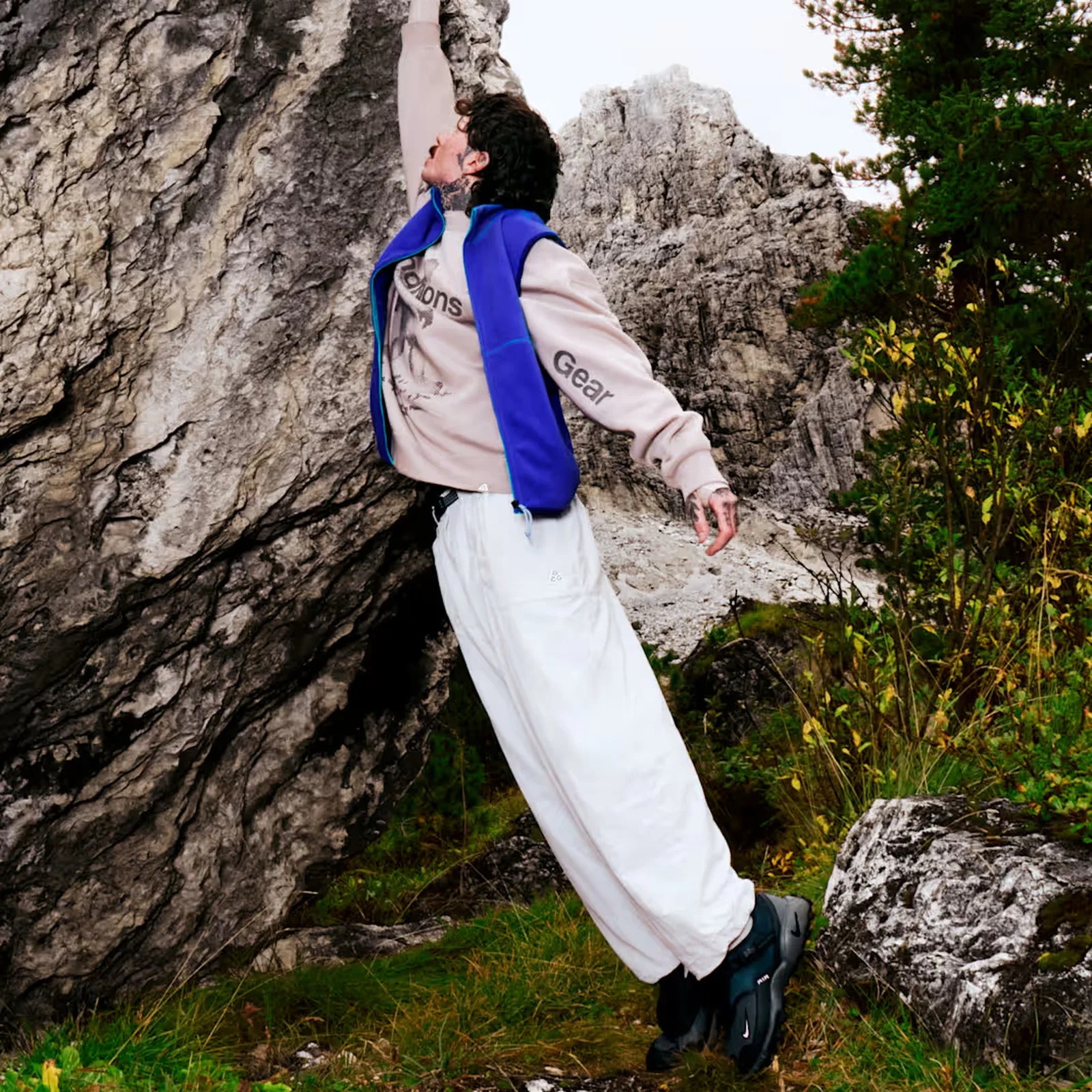 Person wearing a blue vest, beige sweatshirt, white pants, and black shoes climbing a rocky mountain.