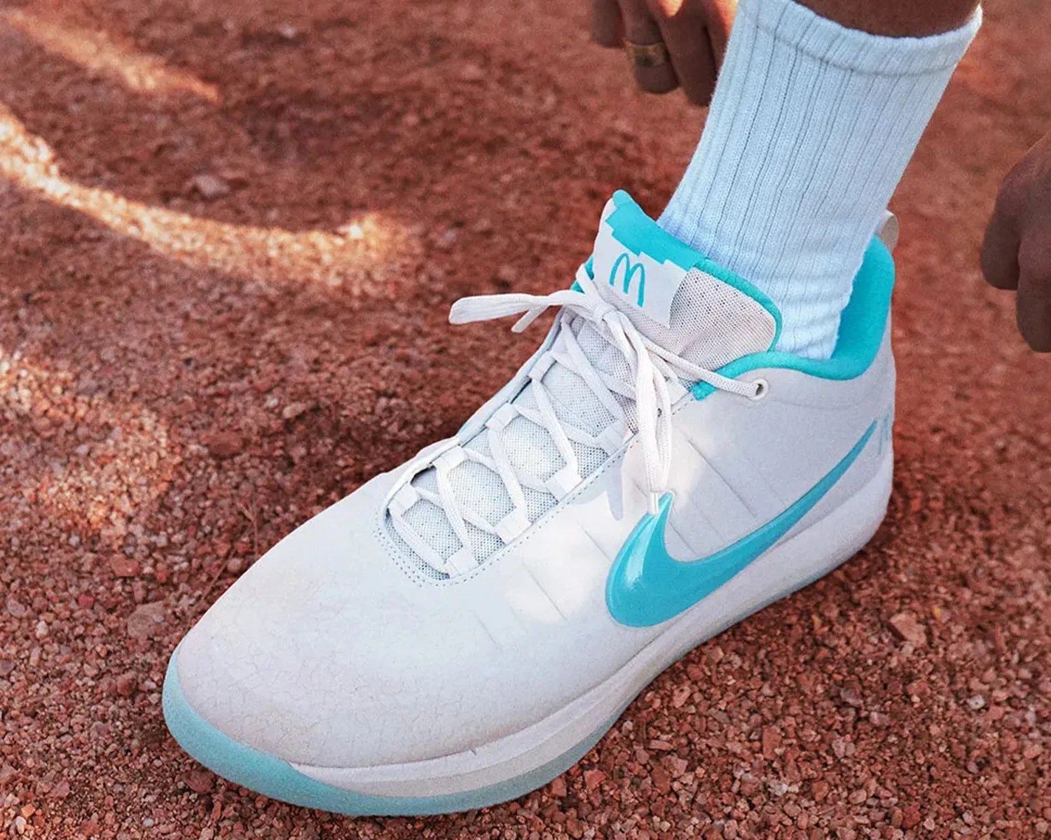 White and turquoise Nike basketball shoe worn with white ribbed socks on a red gravel surface.