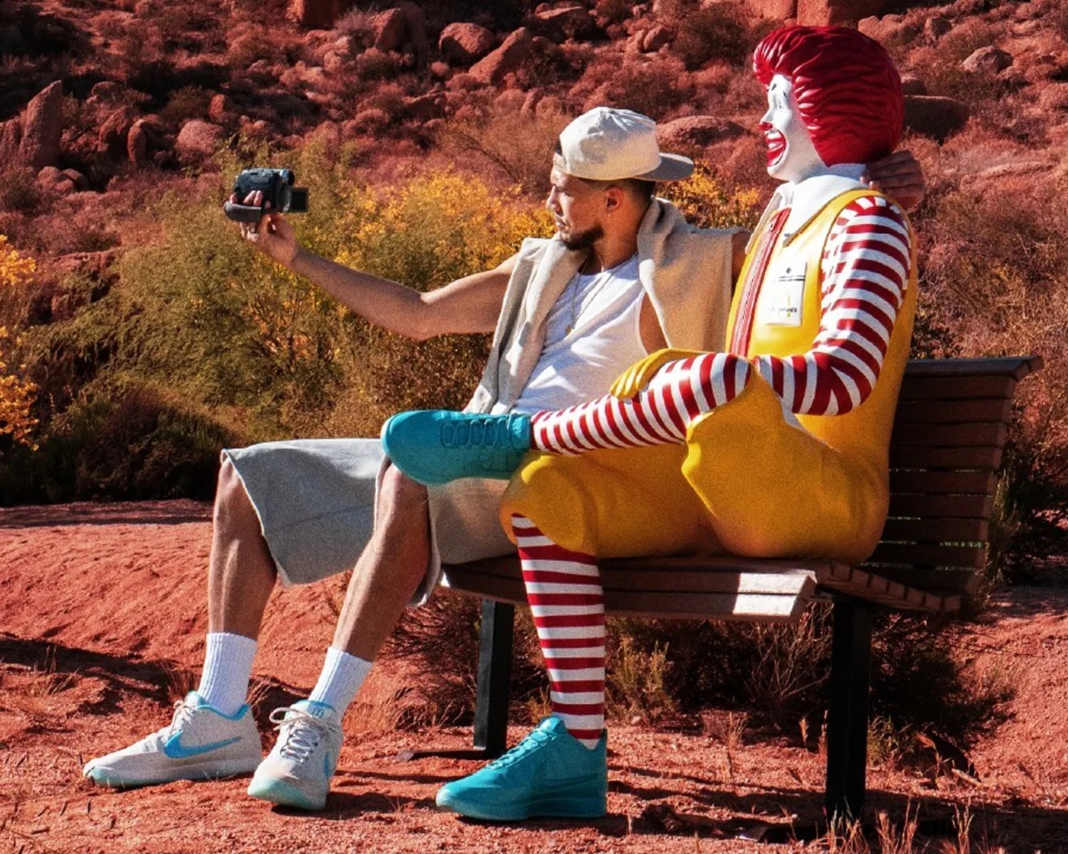 Man in casual clothes and white cap taking a selfie with a Ronald McDonald statue on a bench in a desert setting.