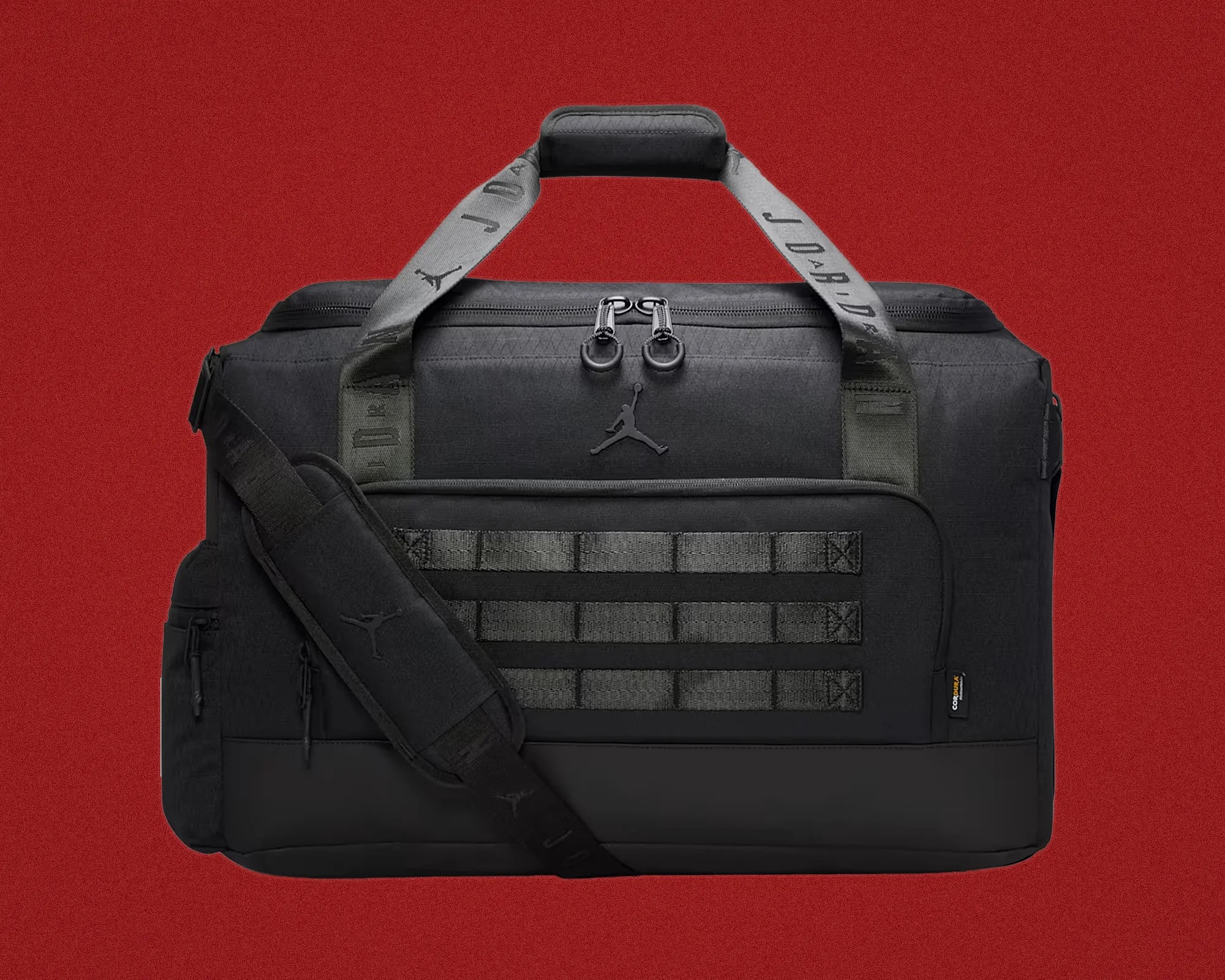 Black Jordan duffel bag with gray branded handles and padded shoulder strap on a red background.
