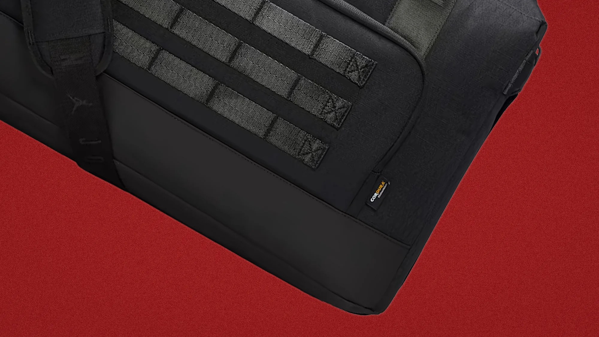 Close-up of a black tactical bag with MOLLE webbing and a small Cordura fabric label on a red background.