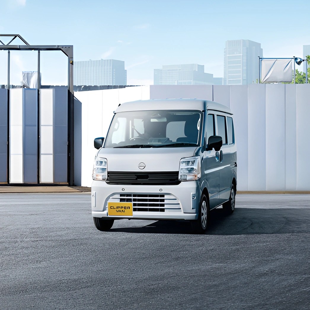 Compact silver van with black grille and yellow license plate parked on asphalt in front of white industrial wall.