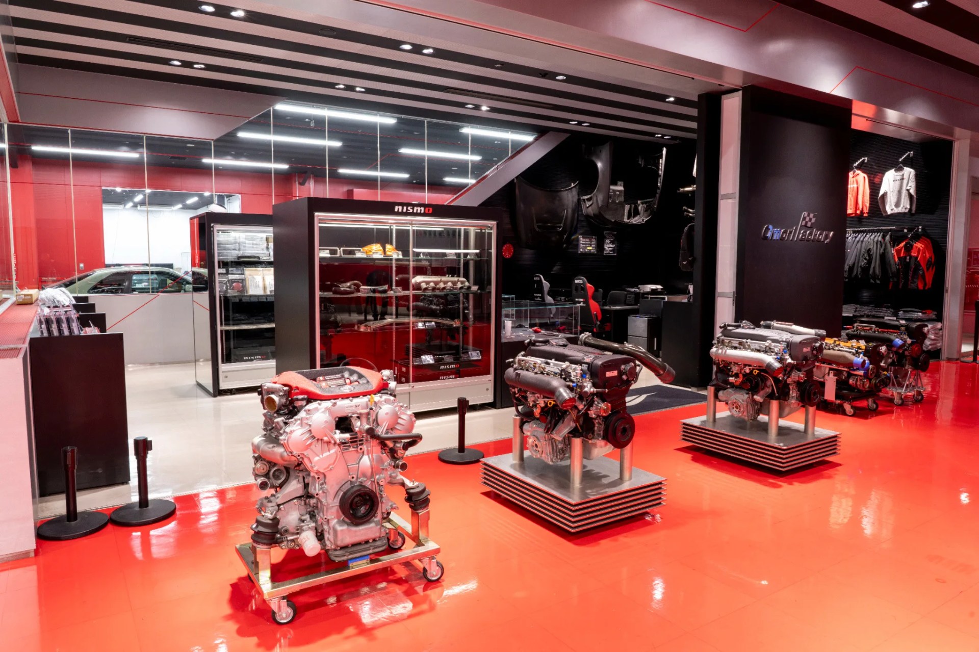 Three car engines displayed on stands inside a showroom with red flooring and a glass display case labeled "nismo.
