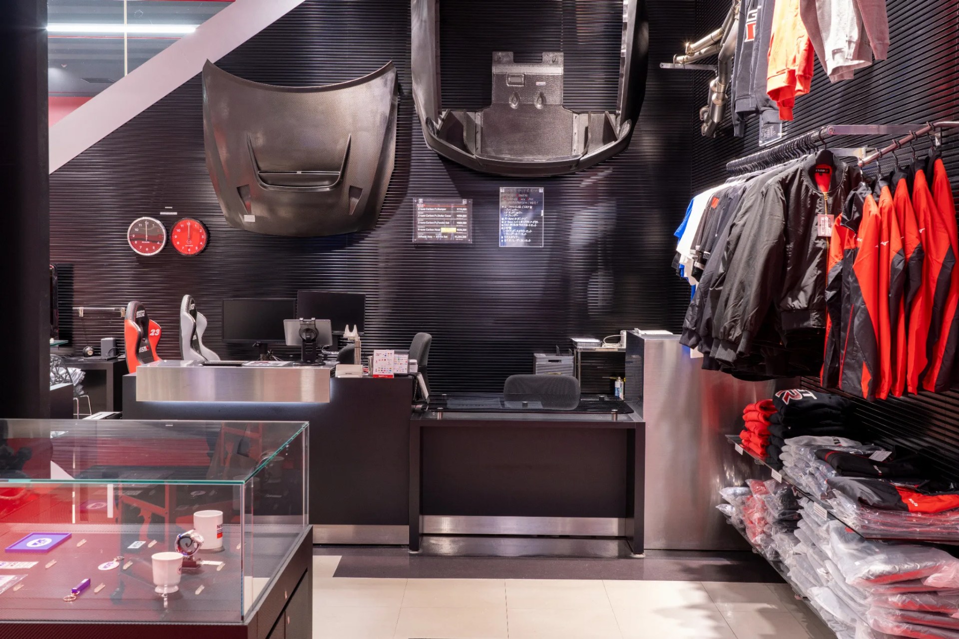 Retail store interior with black walls, carbon fiber car parts mounted, racing seats, and racks of jackets and folded clothes.