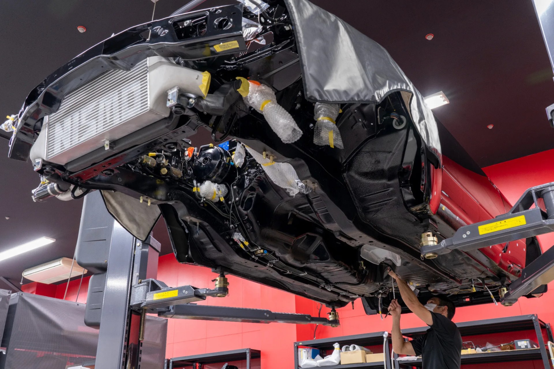 Underneath view of a black car chassis on a lift with bubble-wrapped parts and a person working below in a red-walled garage.