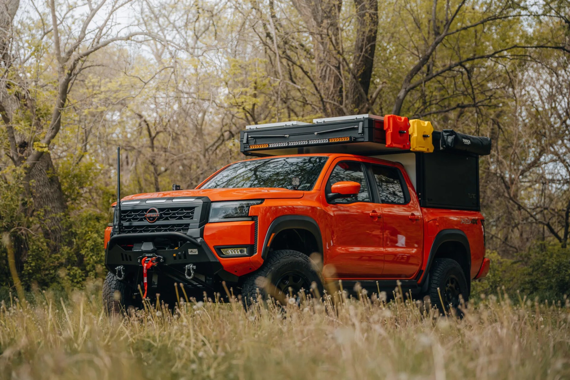 Orange Nissan pickup truck with black roof rack and off-road accessories parked in a grassy area with trees.