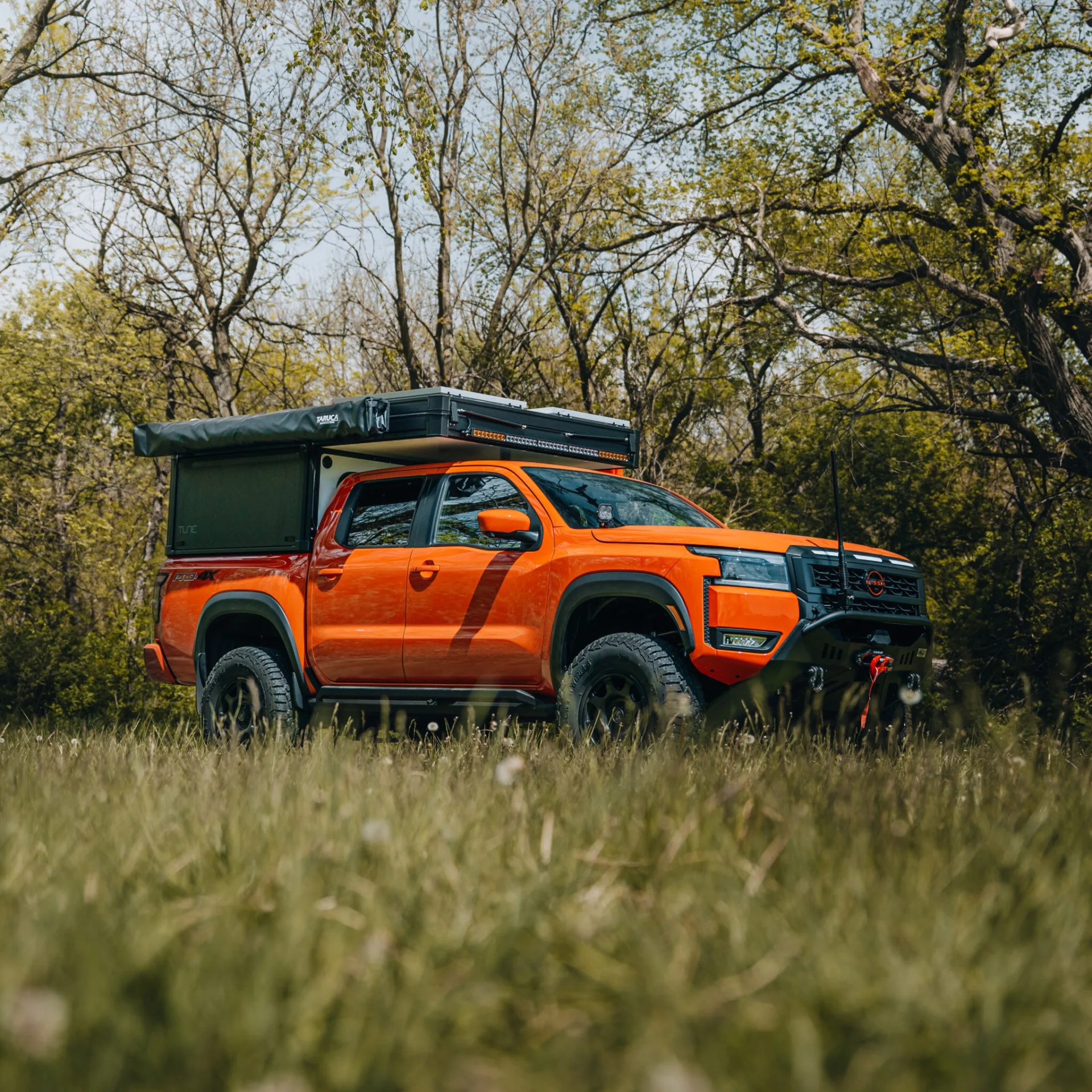 Orange Nissan pickup truck with off-road tires and rooftop tent parked in a grassy area with trees in the background.
