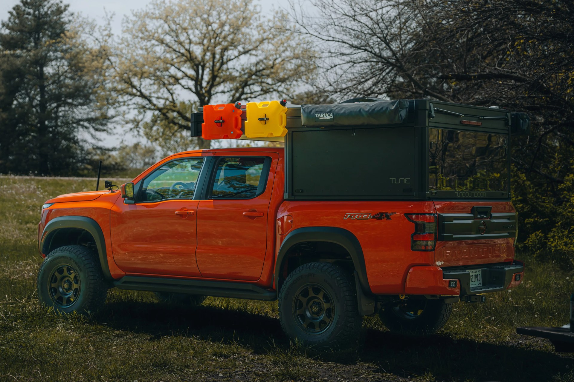 Orange Nissan Frontier Pro-4X pickup truck with black camper shell and mounted red and yellow fuel containers on a grassy field.