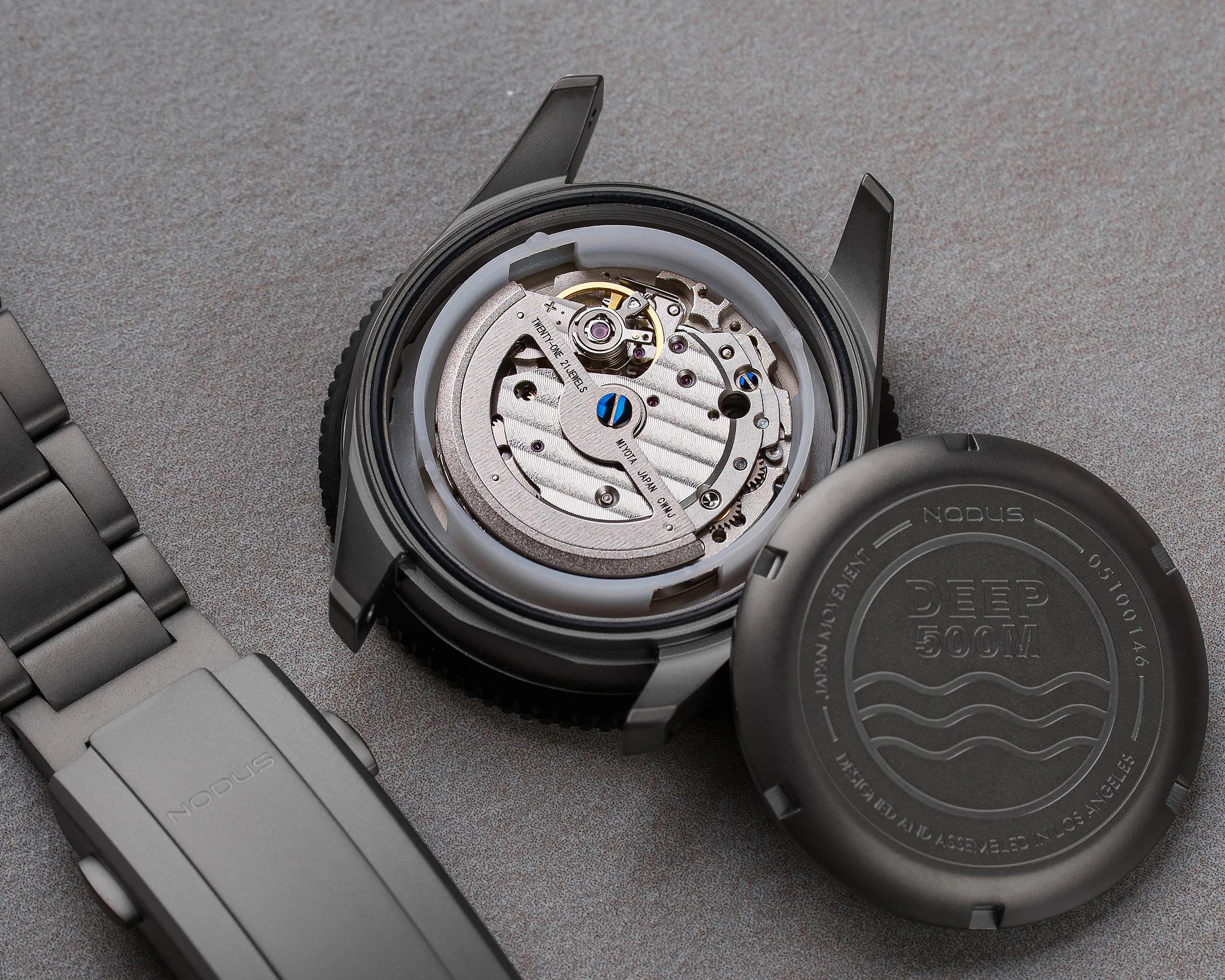 Open black watch case showing silver mechanical movement with blue screws and white plastic ring, next to detached black case back and gray metal bracelet.