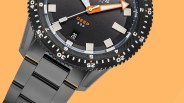 Black dive watch with orange arrow hour hand, white minute markers, and metal link bracelet on orange background.