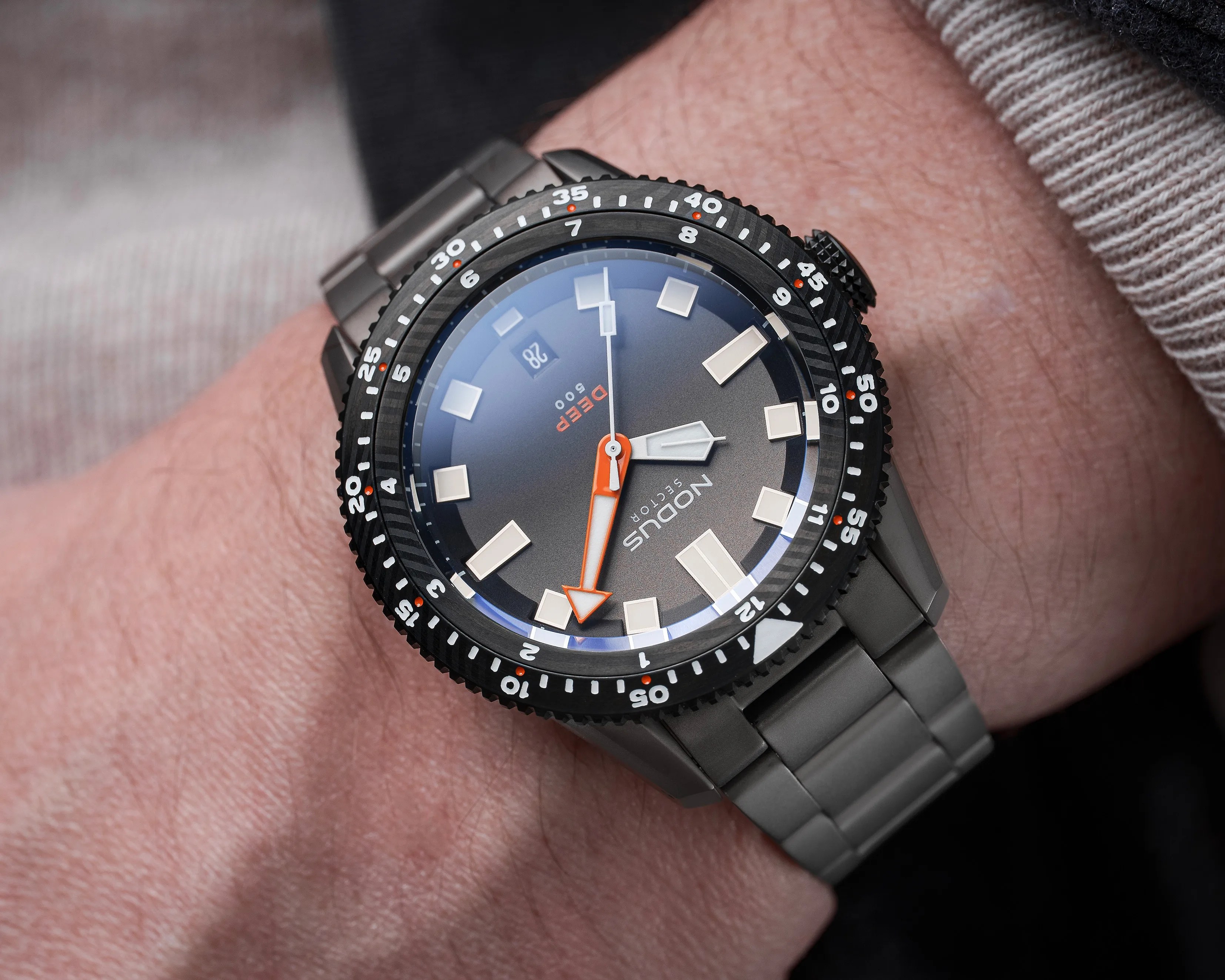 Black dive watch with orange hands and gray metal bracelet on a wrist.