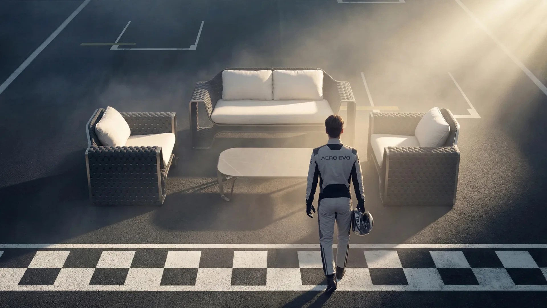 Racing driver in white suit holding helmet walking toward gray woven outdoor sofa set with white cushions on asphalt.