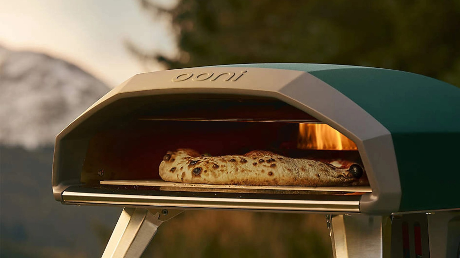 Outdoor green and beige Ooni pizza oven with a pizza baking inside and visible flames.