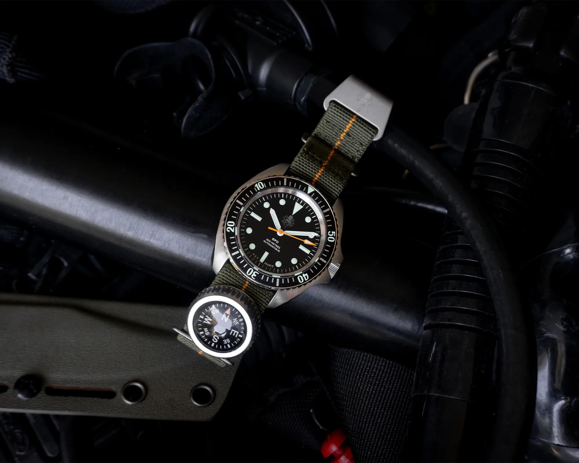 Diver watch with black dial, luminous markers, and green fabric strap with orange stripe, attached to a compass.